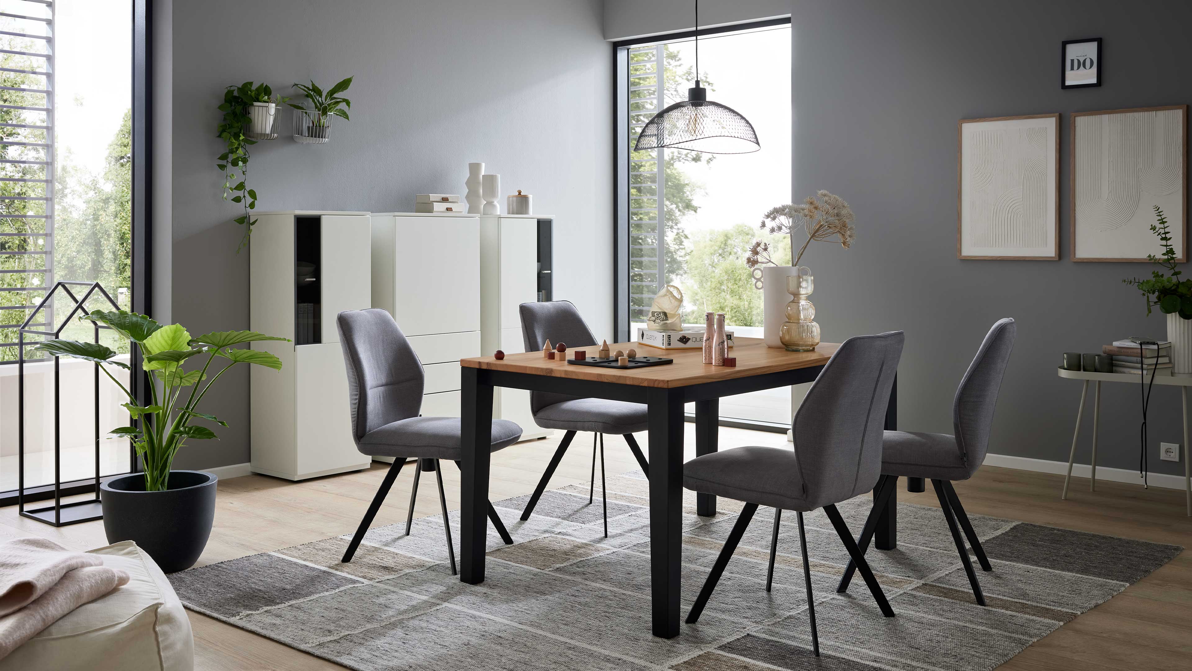 Esszimmertisch mit Holzplatte und schwarzen Beinen, vier graue Polsterstühle, Teppich, weiße Schrankmodule und Hängeleuchte.