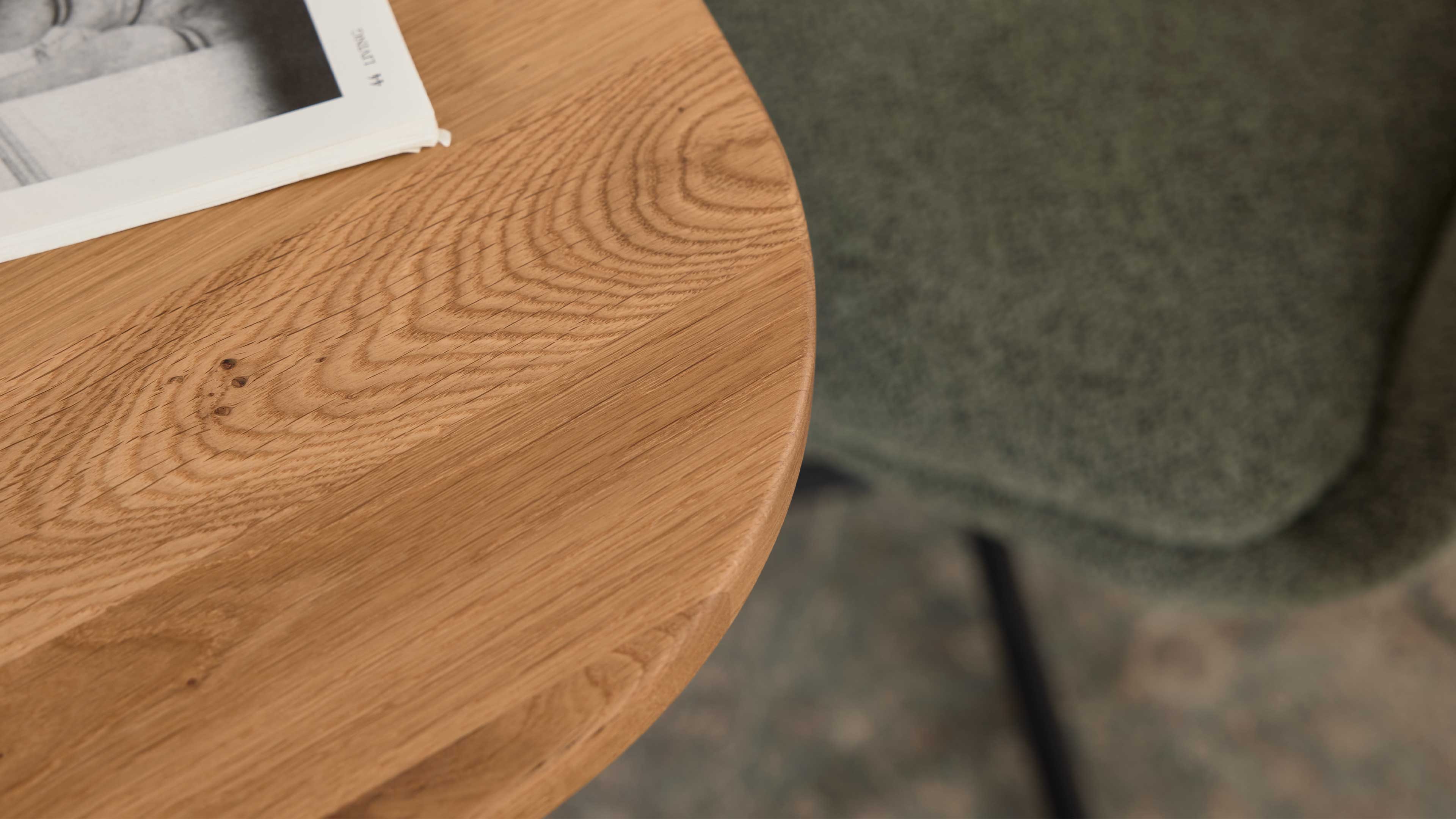 Rundes Holztischblatt mit sichtbarer Maserung, links eine weiße Zeitschrift, rechts ein unscharfer grüner Stoffstuhl mit Bein.