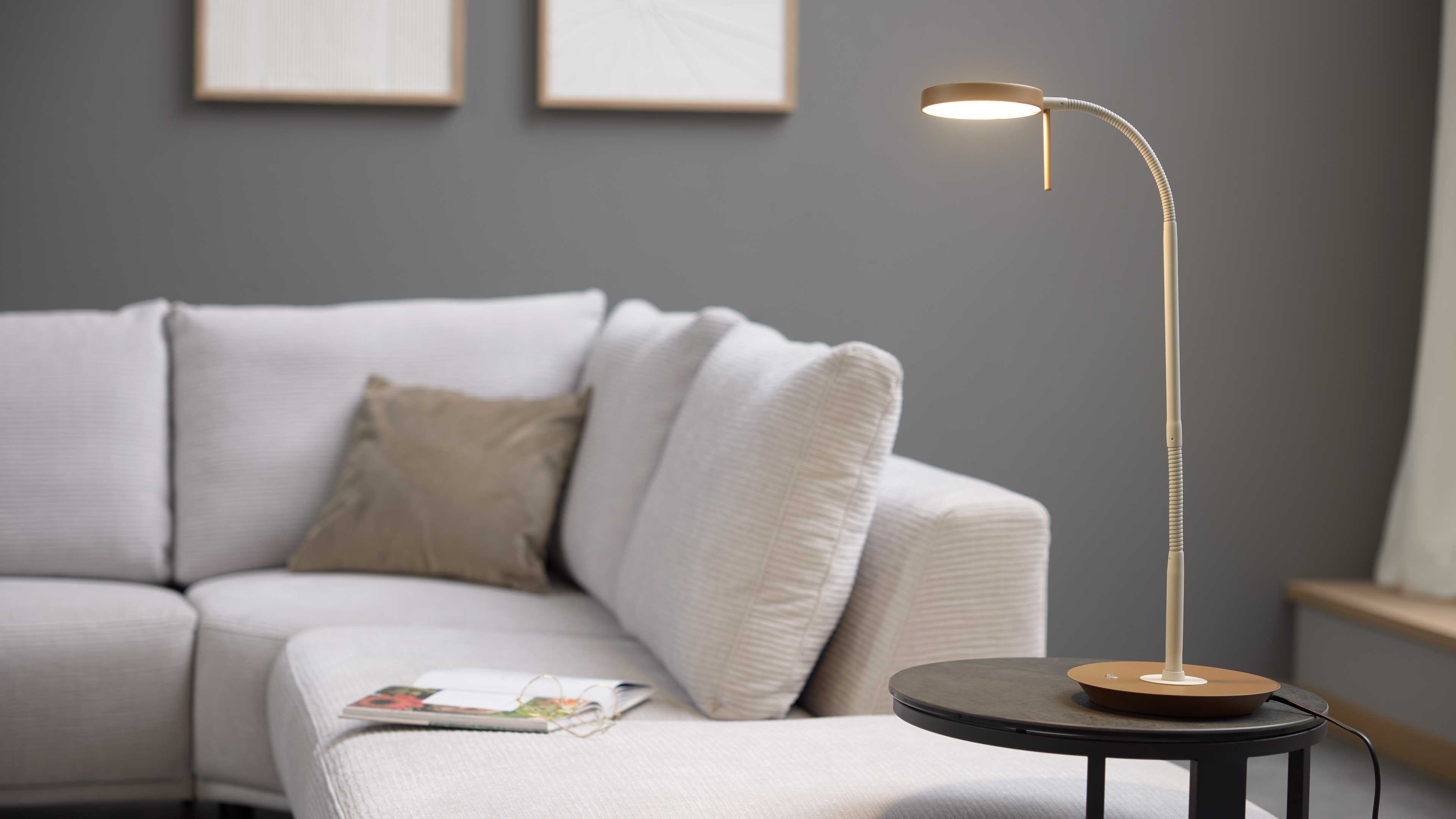 Schlanke beige Tischlampe mit gebogenem Hals auf rundem schwarzen Beistelltisch, davor hellgraues Stoffsofa mit braunem Kissen und Magazin.