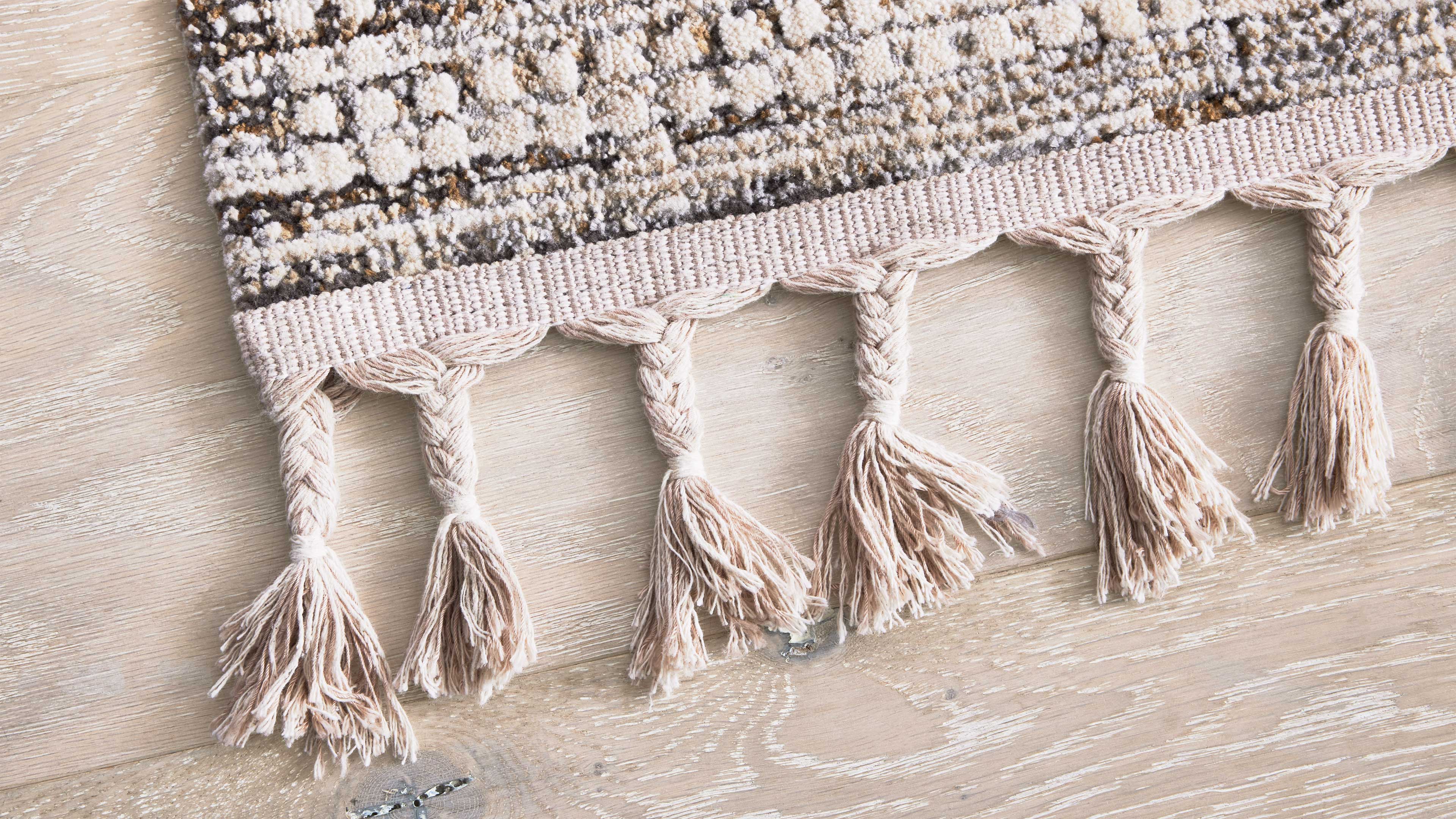 Ecken eines flachgewebten Teppichs mit beigem, gestricktem Saum und sechs geflochtenen Quasten auf hellem Holzfußboden.