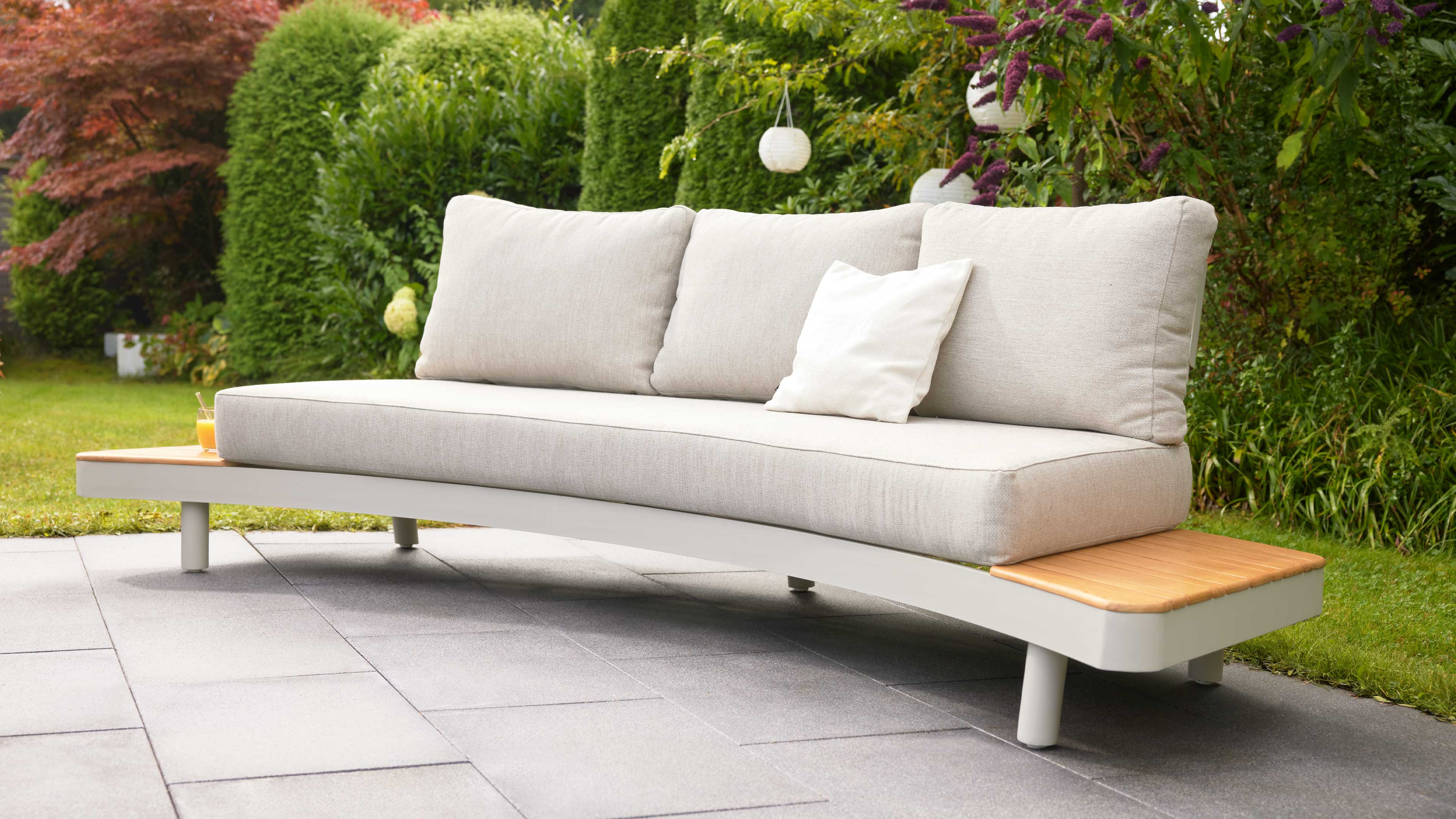Hellbeige Gartenbank mit drei Rückenpolstern und kleinem weißem Kissen, seitlichen Holzablagen auf Terrassenplatten vor Hecke.