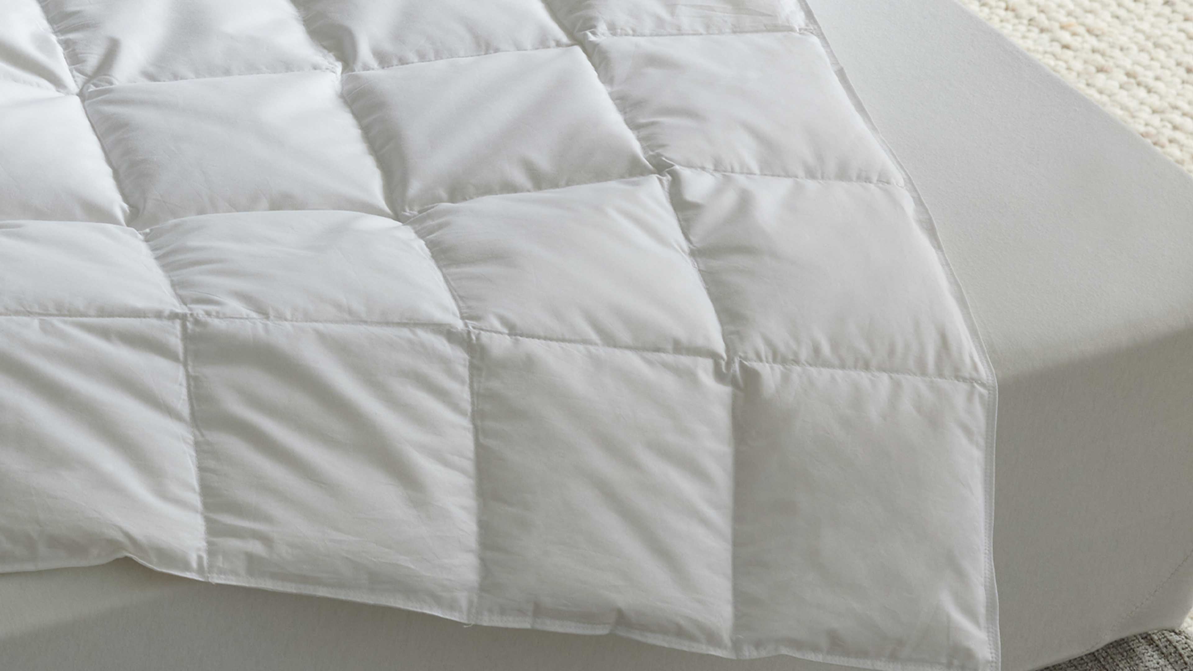 Weiße gesteppte Bettdecke mit quadratischen Kammern liegt auf grauem Spannbettlaken; rechts heller Teppichrand sichtbar.