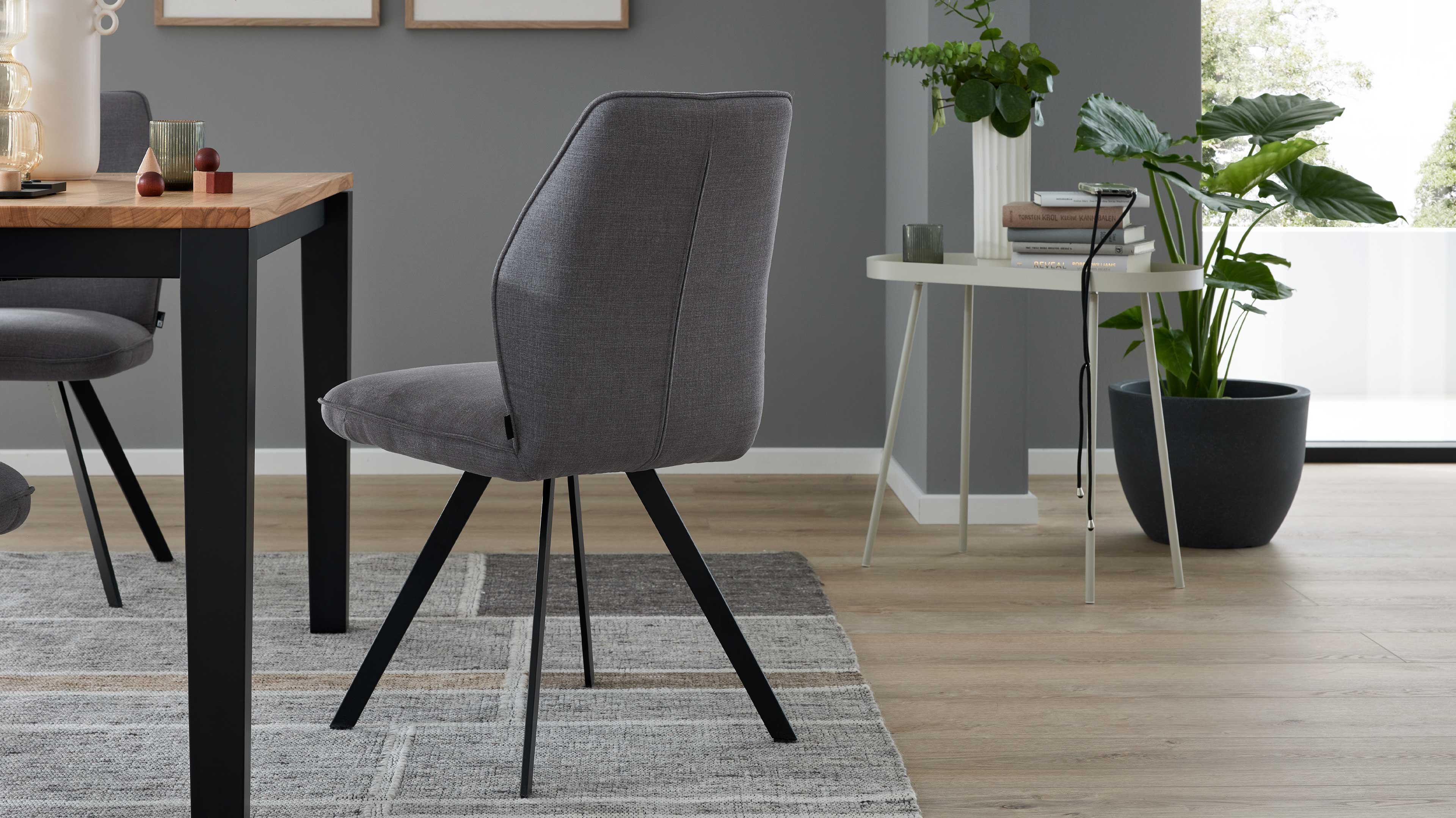Grauer gepolsterter Stuhl mit schwarzen Metallbeinen, neben einem Holztisch mit schwarzem Rand und einem Beistelltisch mit Pflanze.
