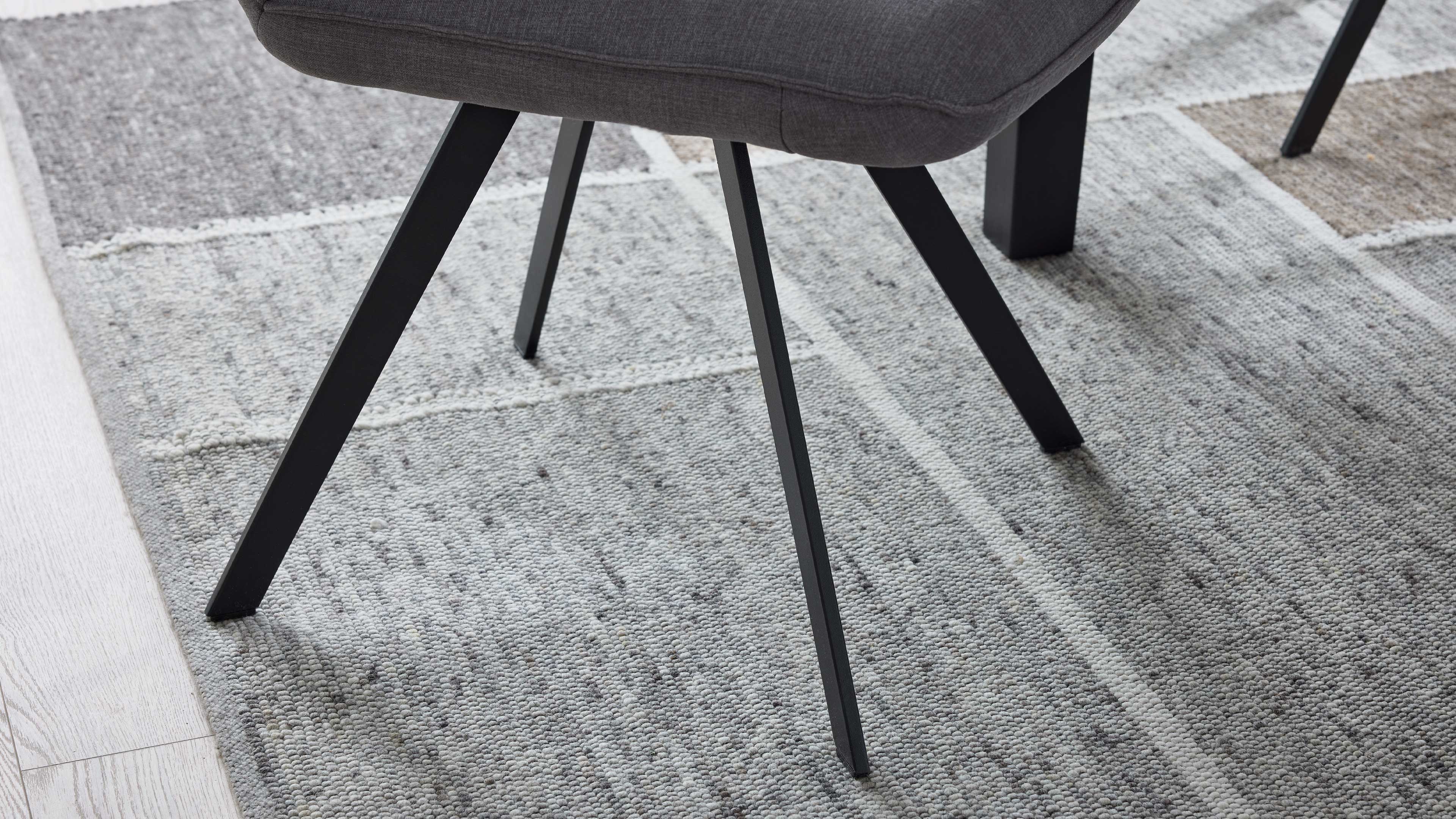 Grau gepolsterte Sitzfläche mit schrägen, schwarzen Metallbeinen auf hellgrauem, strukturiertem Teppich, links Laminatboden sichtbar.