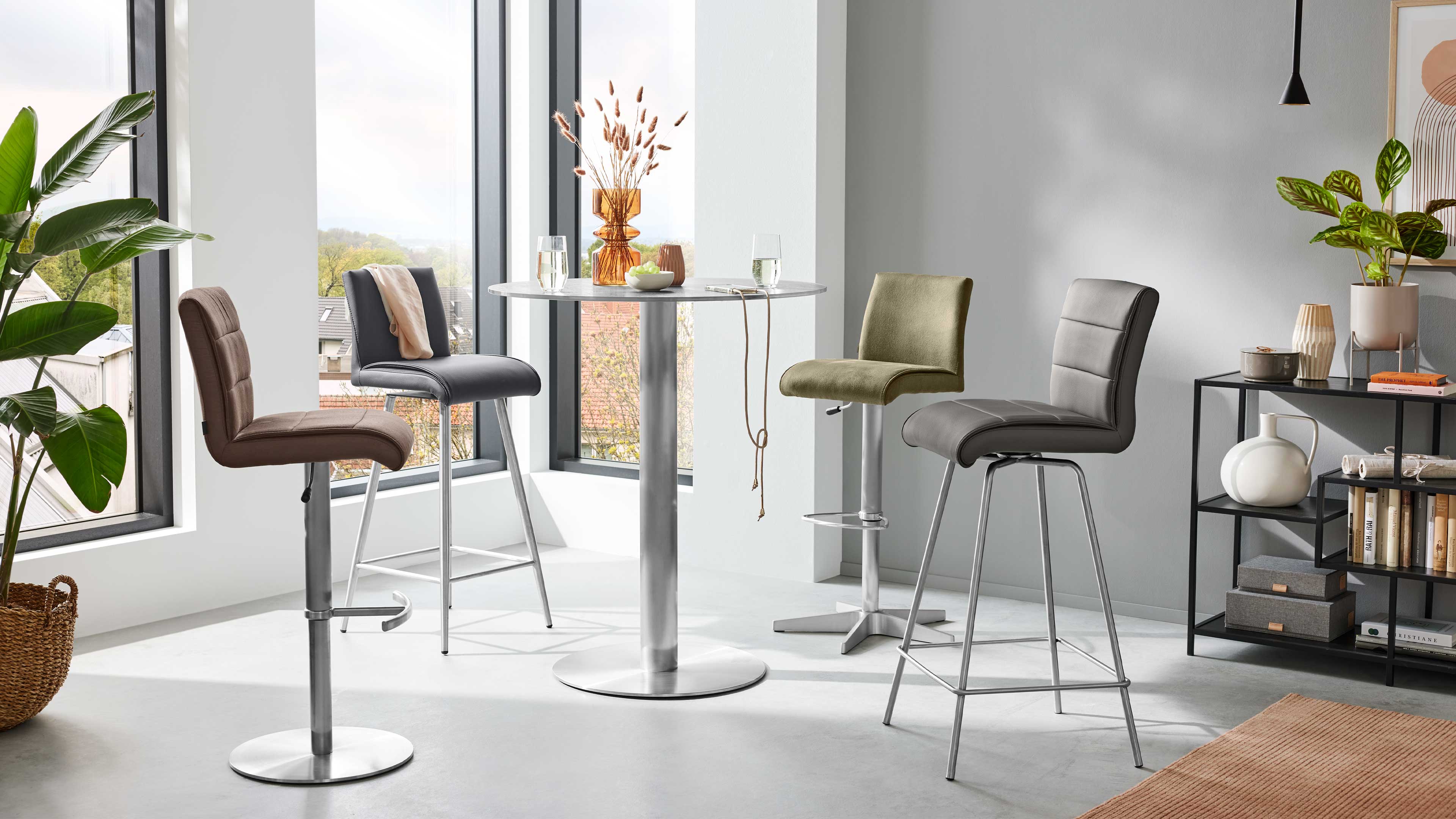 Runder Stehtisch mit Metallfuß und Platte, umgeben von vier gepolsterten Barhockern in verschiedenen Farben vor großen Fenstern.