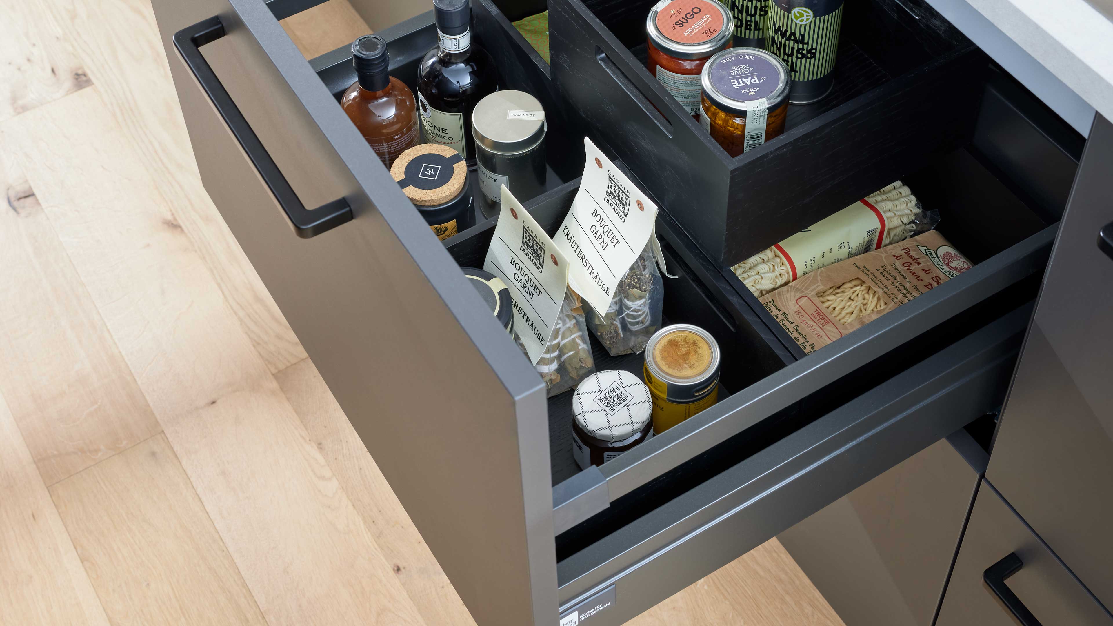 Ausgezogene graue Küchenschublade mit schwarzen Einlegefächern, gefüllt mit Gläsern, Flaschen, Gewürzsträußen und Nudelpäckchen.