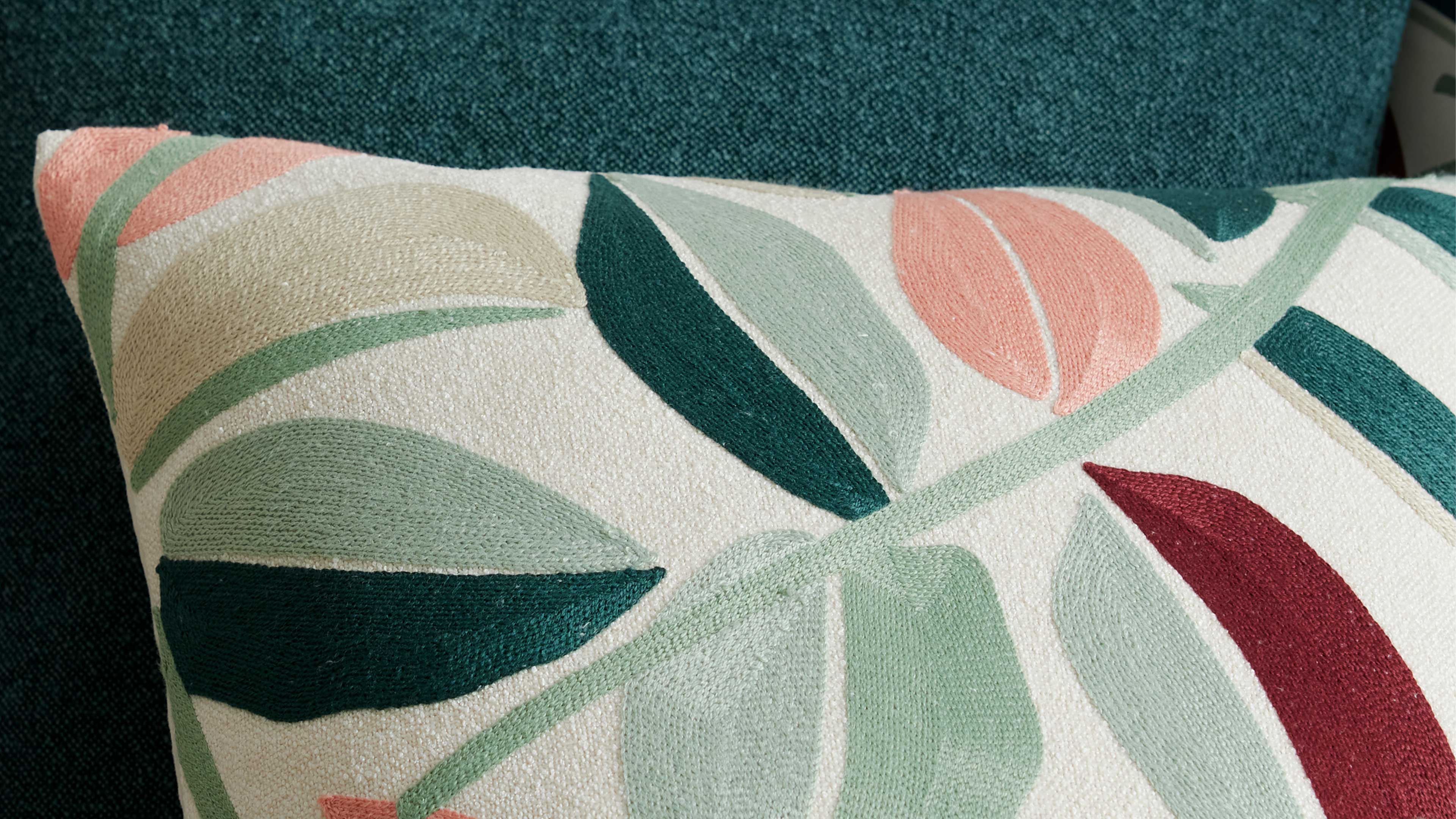 Rechteckiges Kissen mit gestickten Blattmotiven in Grün, Mint, Koralle, Beige und Bordeaux auf hellem Stoff vor petrolfarbener Polsterfläche.