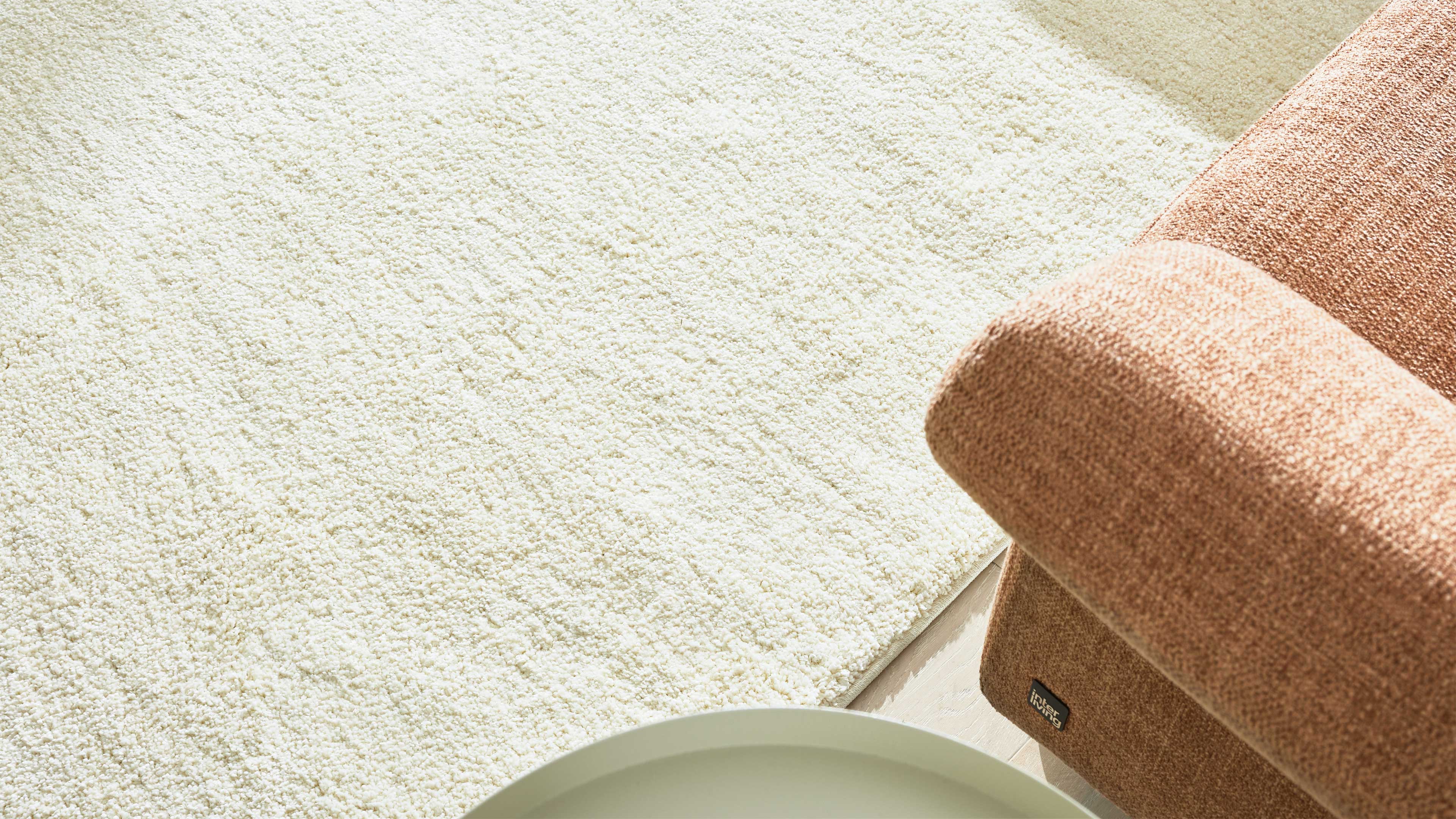 Rechts ein rostfarbenes Sofaarmteil mit grobem Stoff, links ein cremefarbener, dichter Teppich und unten eine runde Tischkante.