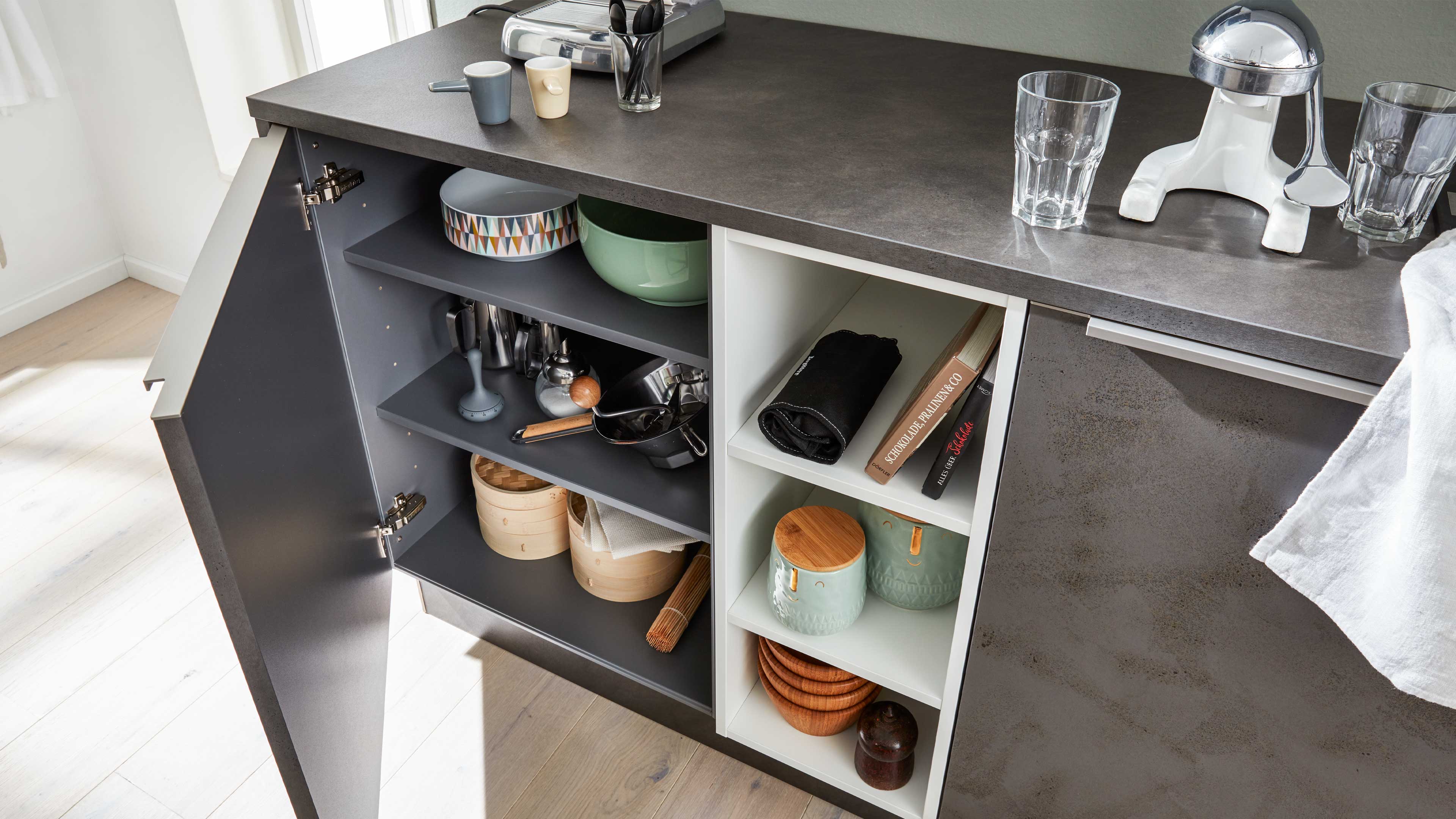 Offener Küchenschrank mit anthrazitfarbener Front und dunkler steiniger Arbeitsfläche, innen links graue Regalböden und rechts weiße Fächer.