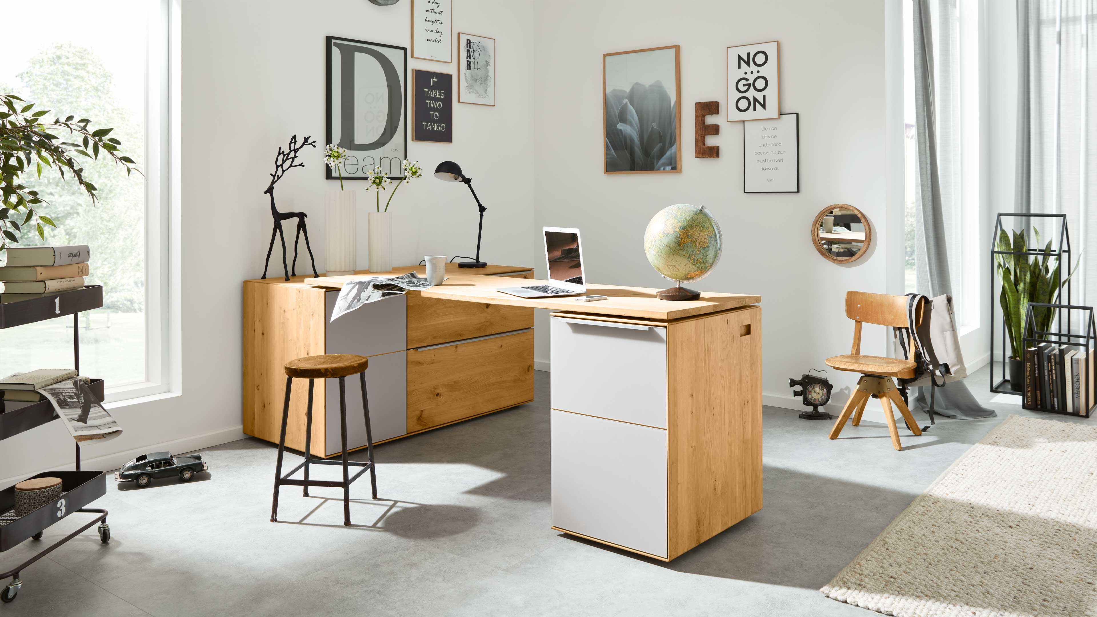 L-förmiger Holzschreibtisch mit weißen Schubladen, Laptop und Globus in hellem Raum, dazu Hocker, Holzstuhl, Pflanze und Wandbilder.