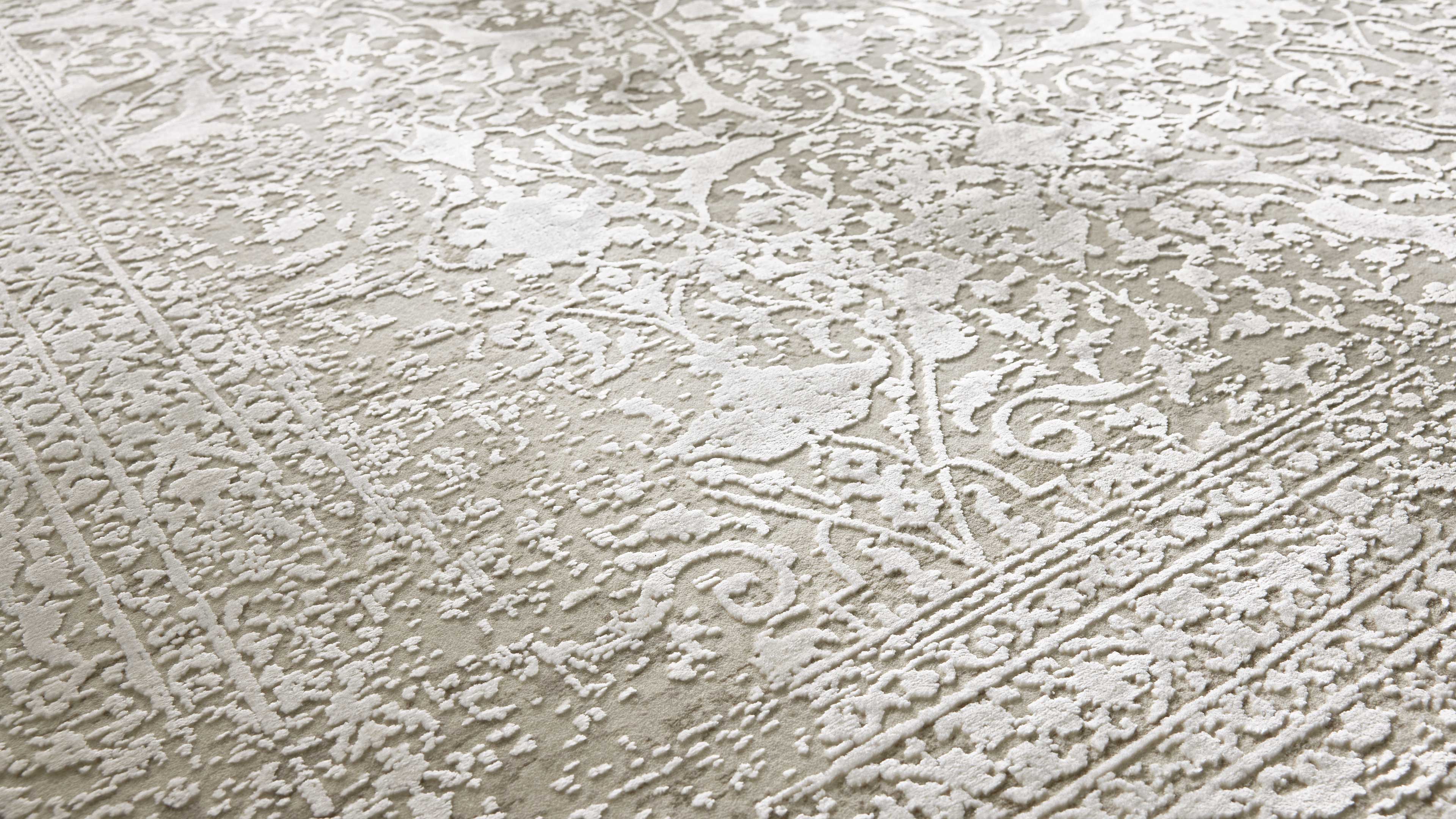 Teppich mit hellbeigem Grund und erhöhtem, floralen Ornamentmuster in cremeweiß, sichtbare Bordüren und feine Details.