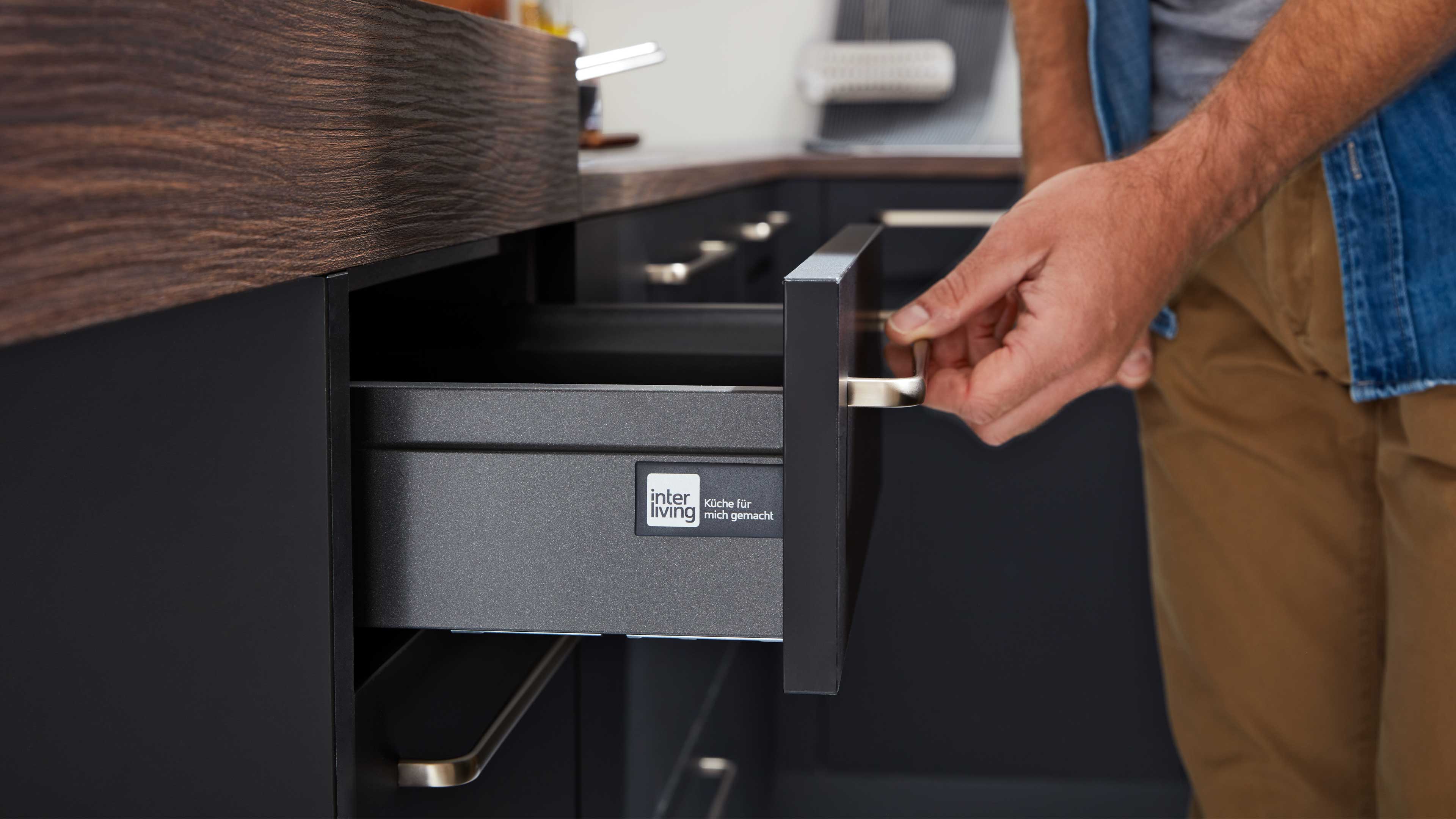 Schwarzer Küchenschubkasten wird von einer Hand geöffnet, silberner Griff unter Holzarbeitsplatte, 'interliving'-Etikett sichtbar.