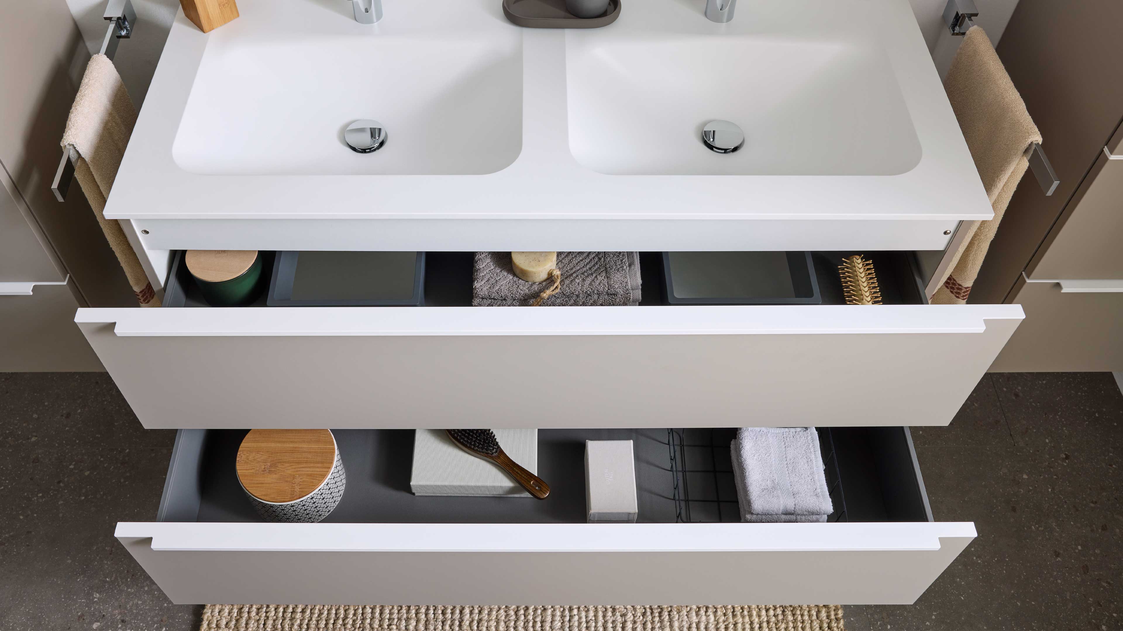 Weißes Doppelwaschbecken auf beigem Unterschrank mit zwei offenen Schubladen, darin Handtuch, Seife, Bürste und Behälter.