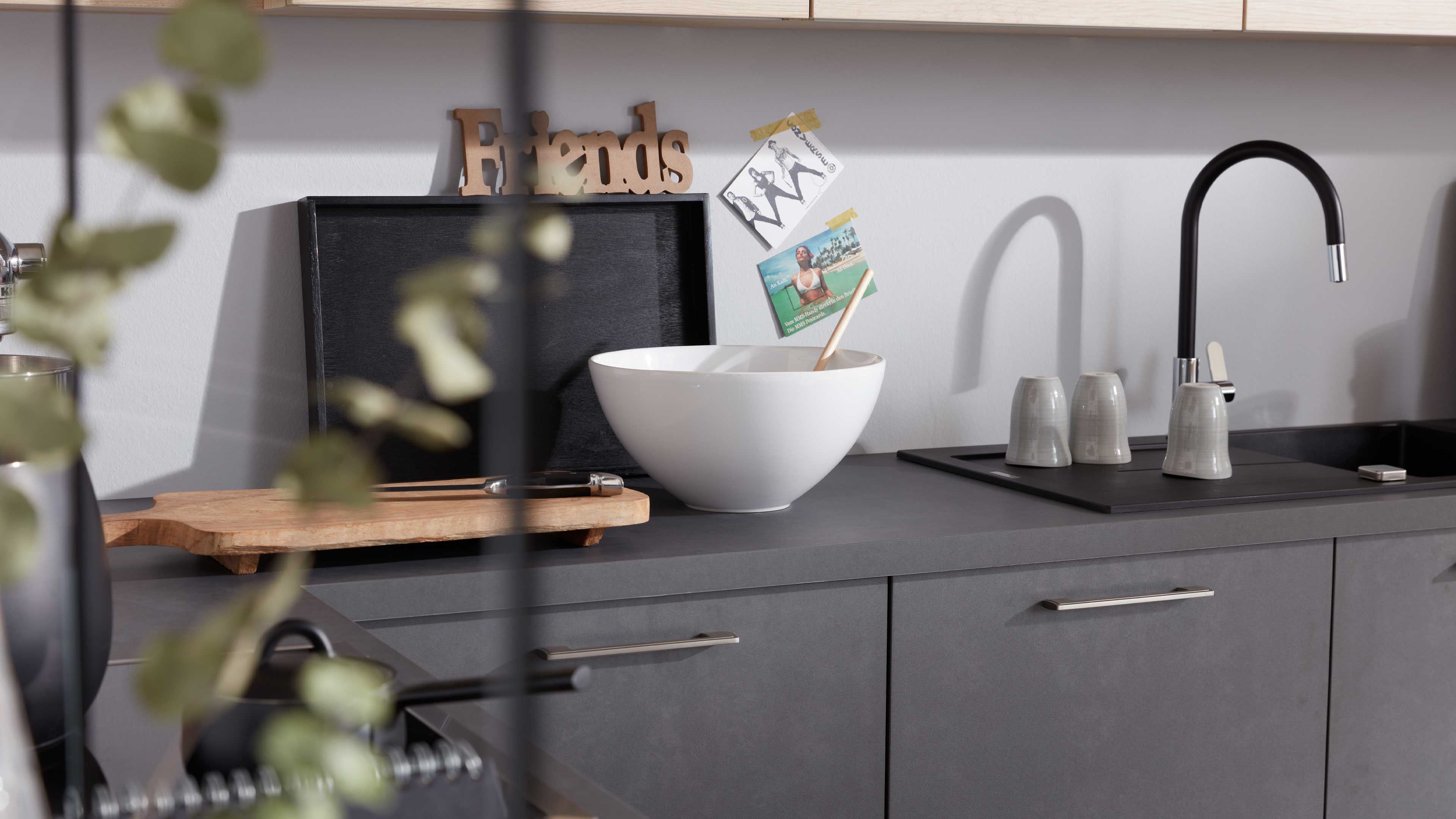 Küchenarbeitsplatte mit dunkelgrauen Schrankfronten, weißer Rührschüssel, Holzbrett, schwarzer Armatur mit hohem Bogen und drei grauen Tassen.