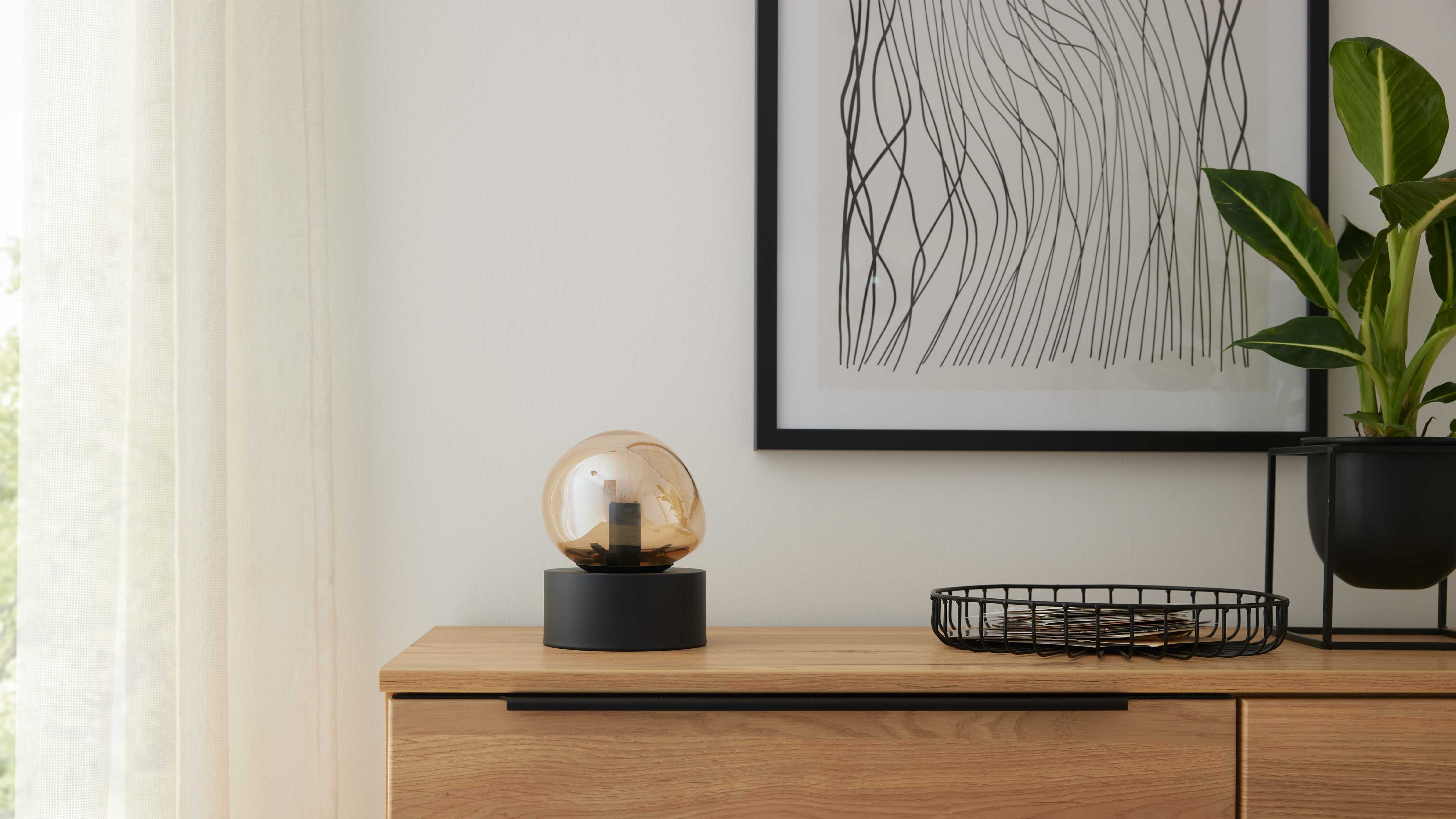Hölzernes Sideboard mit schwarzer Sockellampe mit bernsteinfarbener Glaskugel, Drahtkorb, Pflanze und gerahmter Linienzeichnung an der Wand.