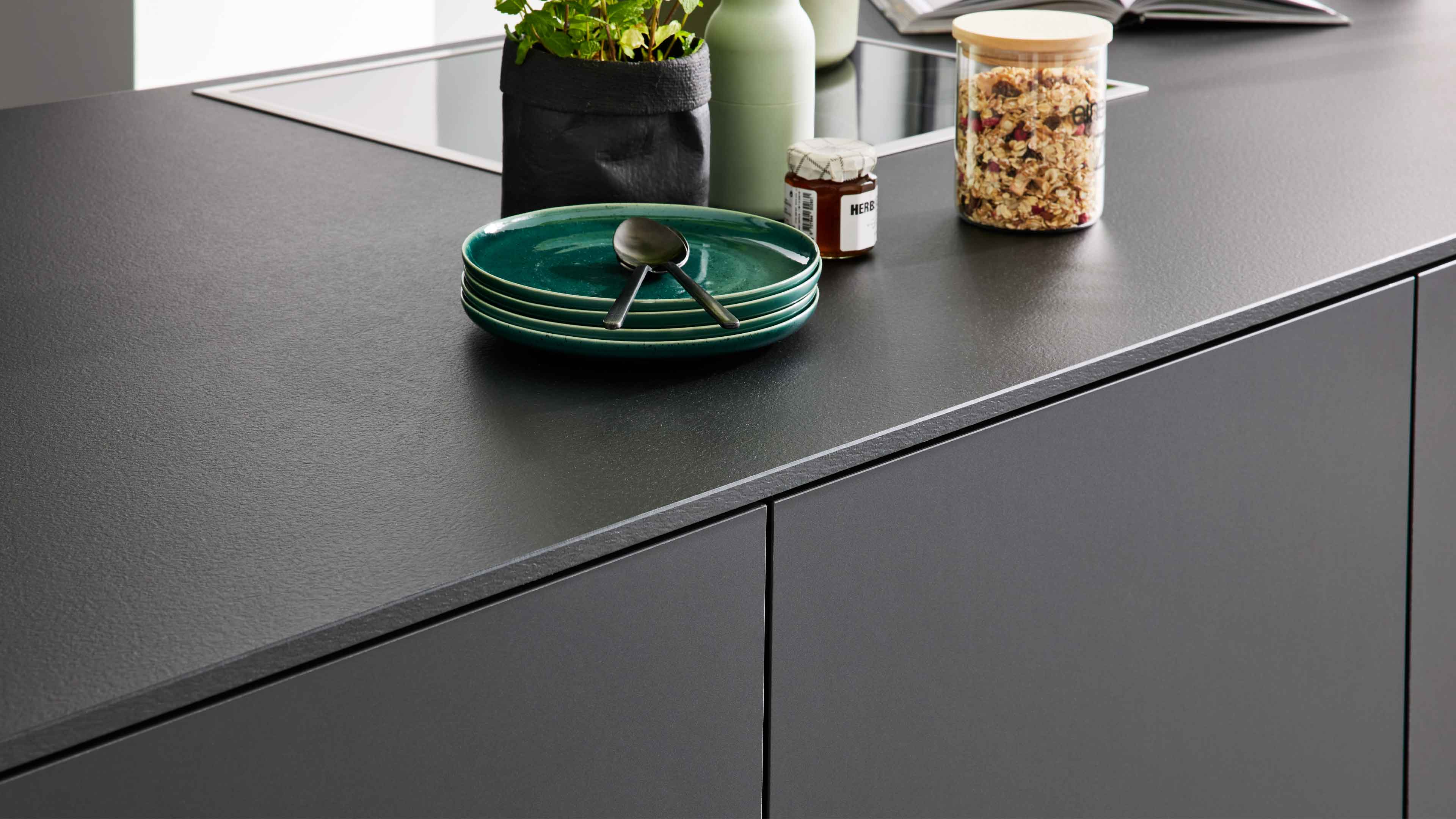 Dunkle Küchenarbeitsplatte mit gestapelten grünen Tellern und zwei Löffeln, schwarzem Pflanzenbeutel, kleinem Glas und Müsli im Glas.