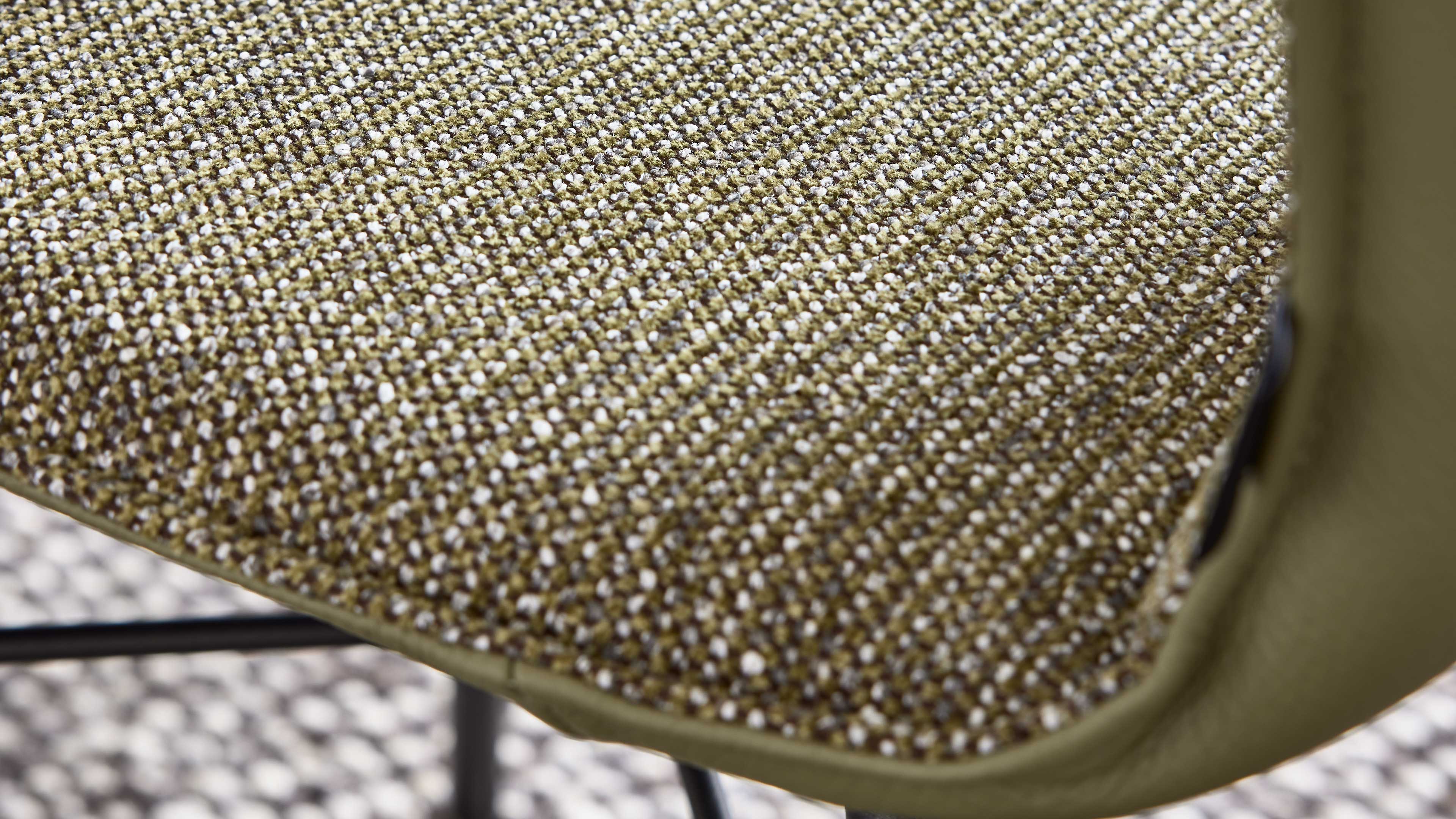 Nahaufnahme einer gepolsterten Sitzfläche mit olivgrün-weiß melierter, grob gewebter Stoffstruktur und sichtbarem schwarzen Metallgestell.