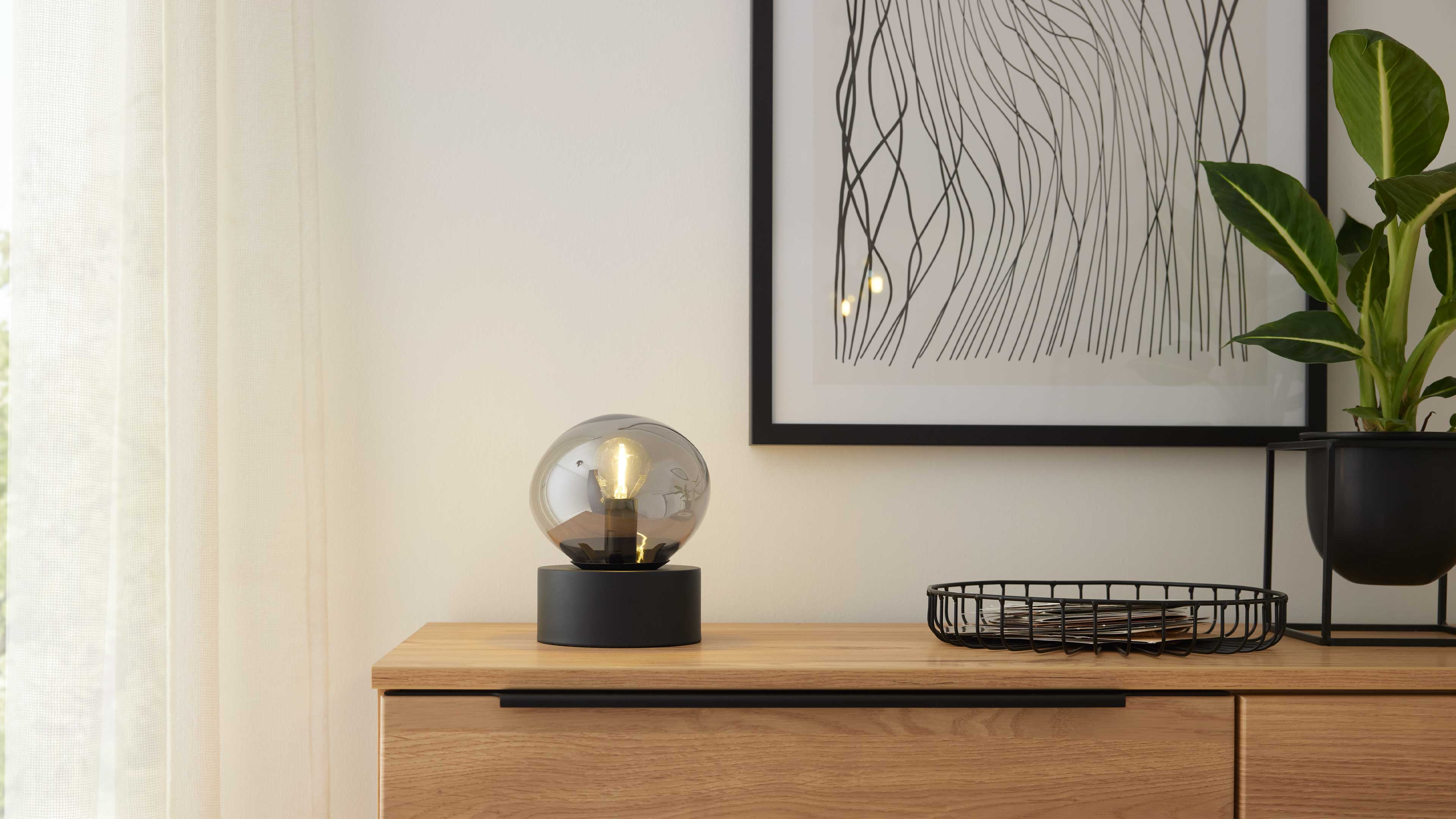 Holzkommode mit runder Rauchglas-Tischleuchte auf schwarzem Sockel, Drahtkorb, Pflanzenübertopf und gerahmtem Linienbild.