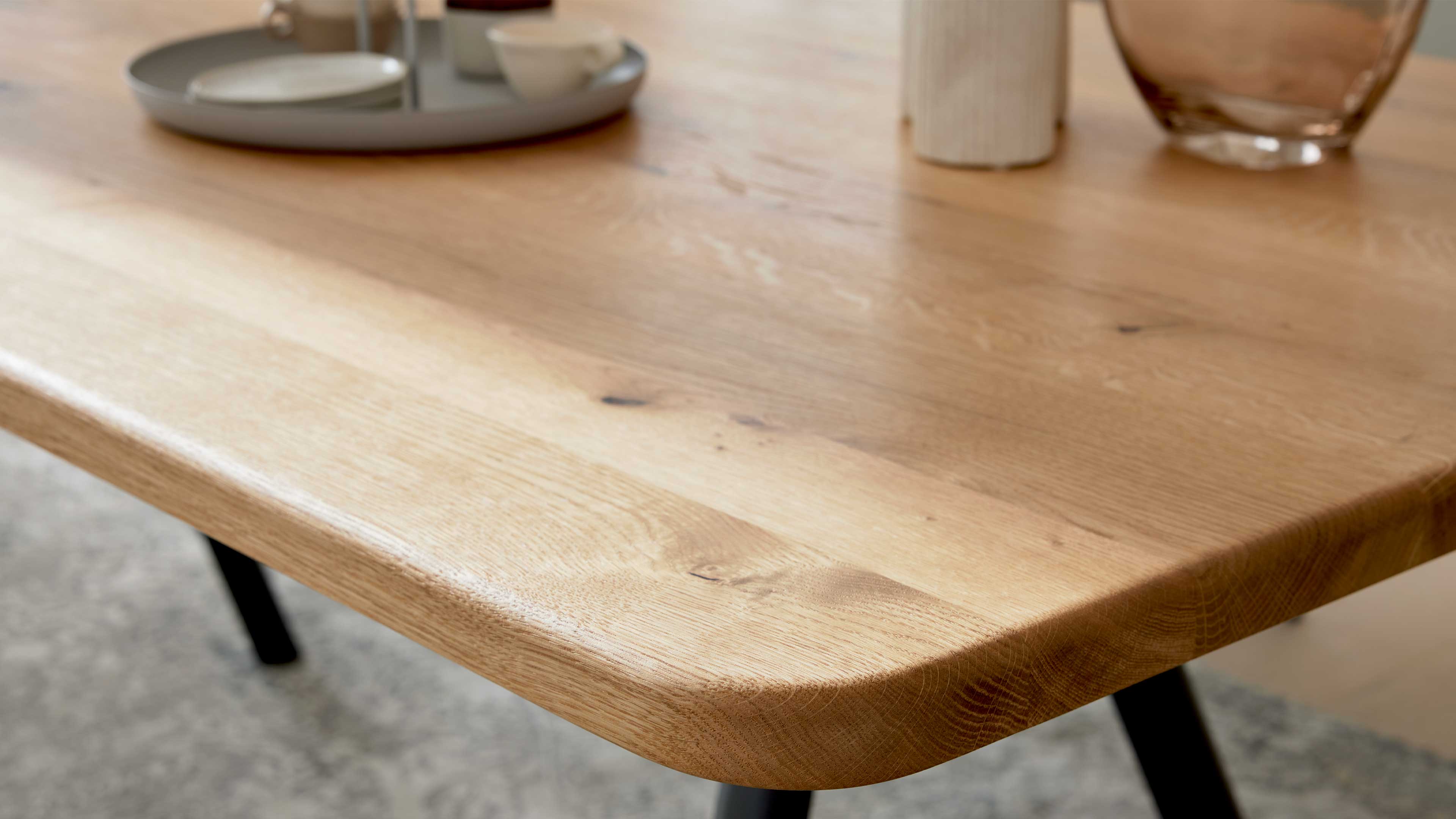 Tischplatte aus hellem Holz mit sichtbarer Maserung und abgerundeter Kante, schwarze Beine sowie runde Schale und Glasvase im Hintergrund.