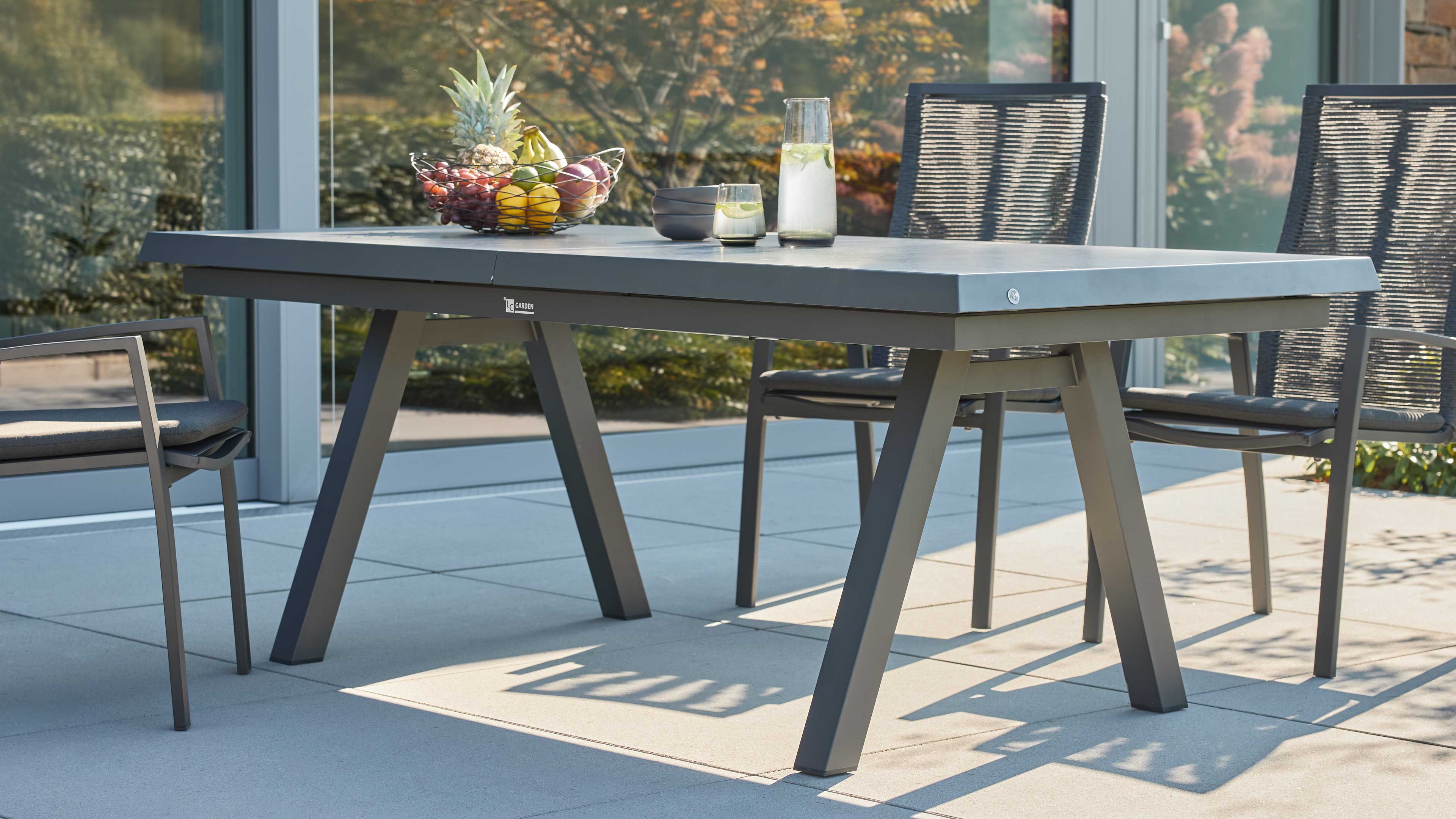 Grau lackierter rechteckiger Gartentisch mit abgeschrägten Beinen, umgeben von Stühlen, mit Obstkorb und Karaffe.
