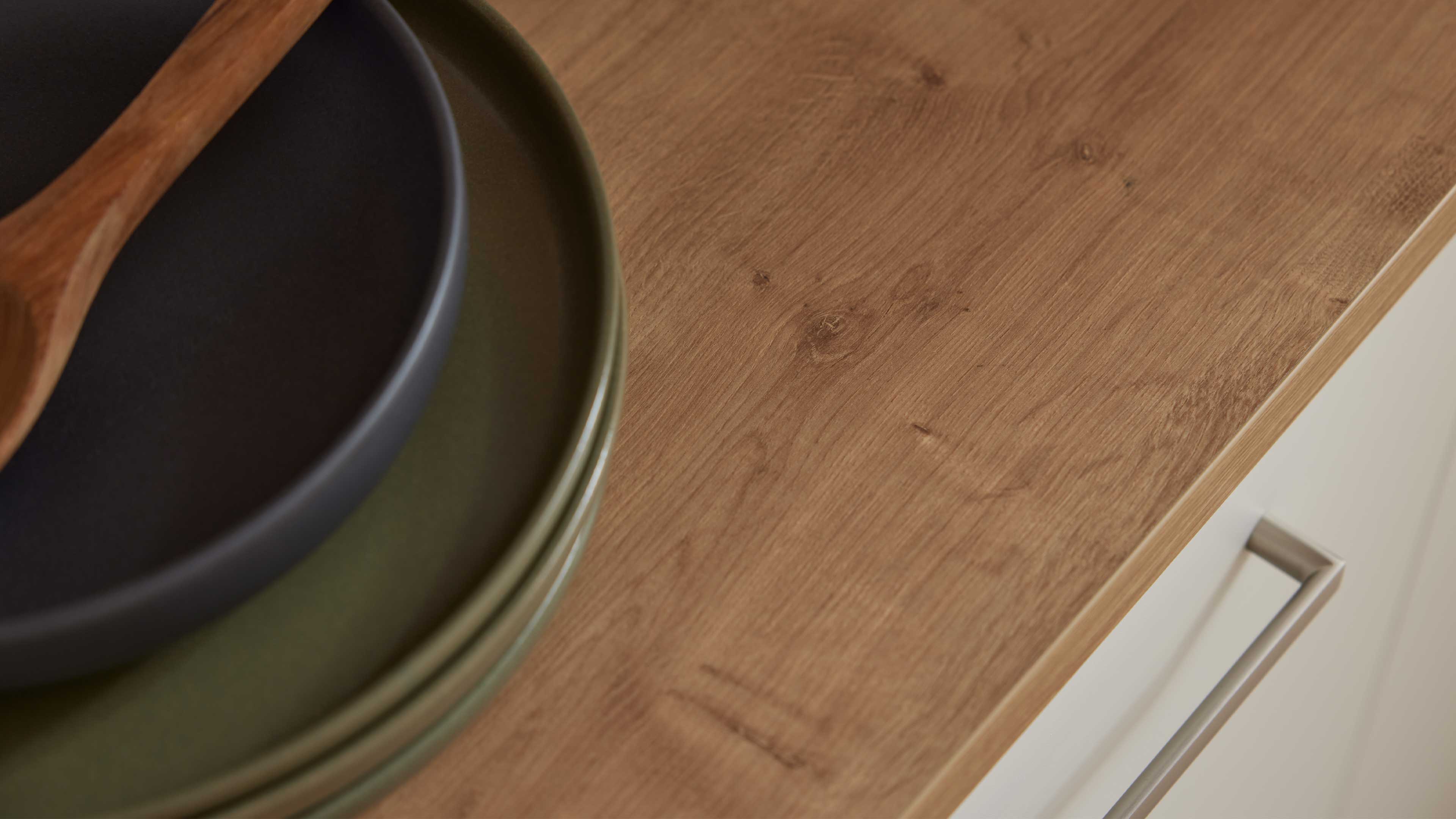 Holzarbeitsplatte mit sichtbarer Maserung und Kante, links gestapelte grüne und schwarze Teller sowie ein Holzlöffel.