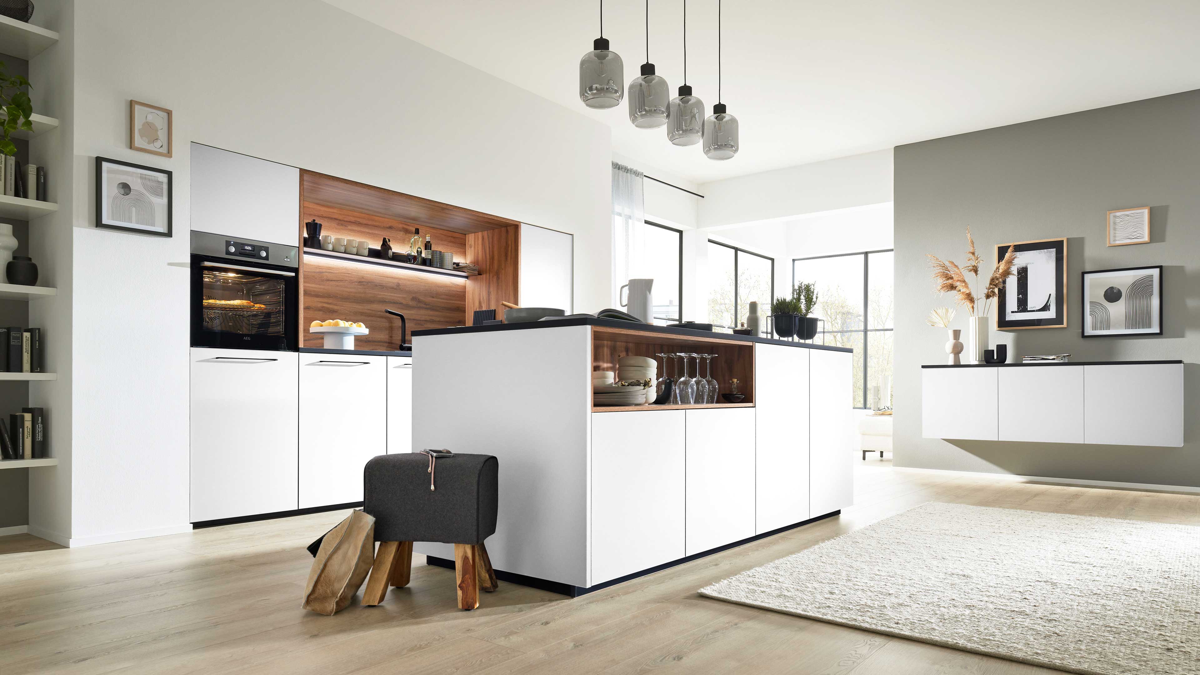 Weiße Kücheninsel mit offenem Holzfach voller Gläser und Geschirr, daneben Hochschränke mit eingebautem Ofen und grauem Hocker.