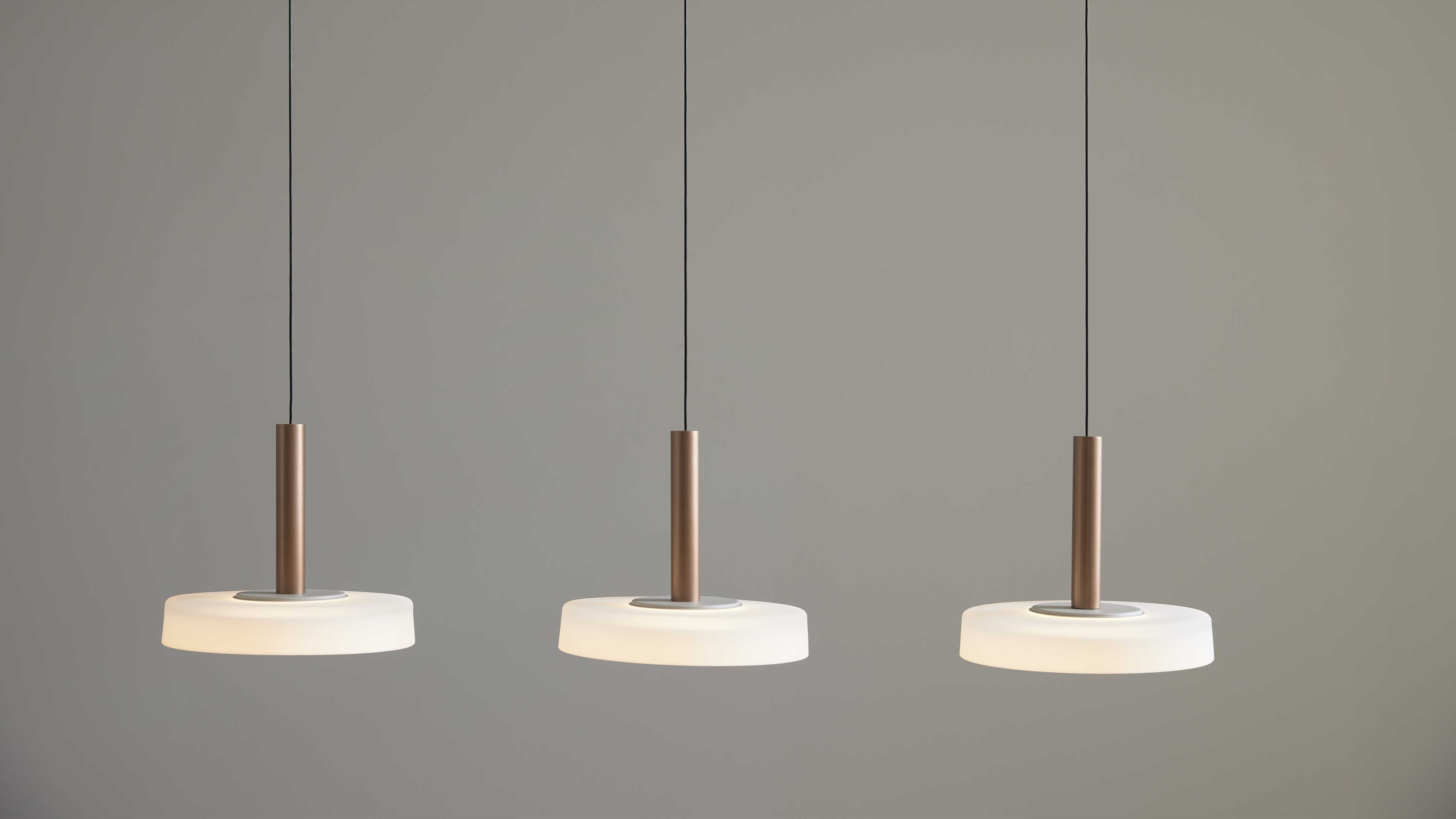 Drei hängende Deckenlampen mit braunen zylindrischen Stangen und flachen weißen Diffusoren, an schwarzen Kabeln vor grauer Wand.