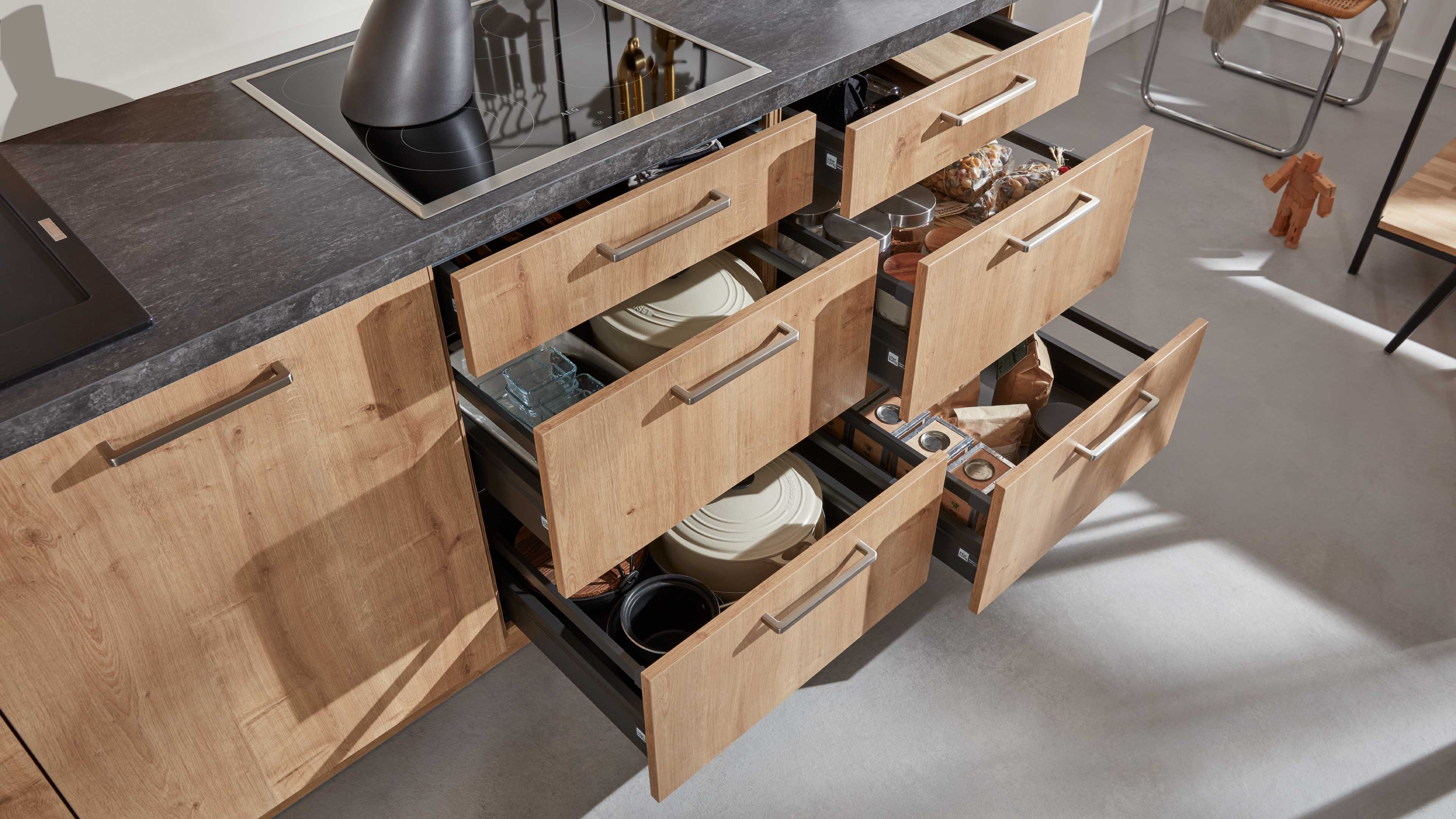 Küchenunterschränke mit hellen Holzfronten und Metallgriffen, dunkler Arbeitsplatte und geöffneten Schubladen mit Töpfen und Vorratsdosen.