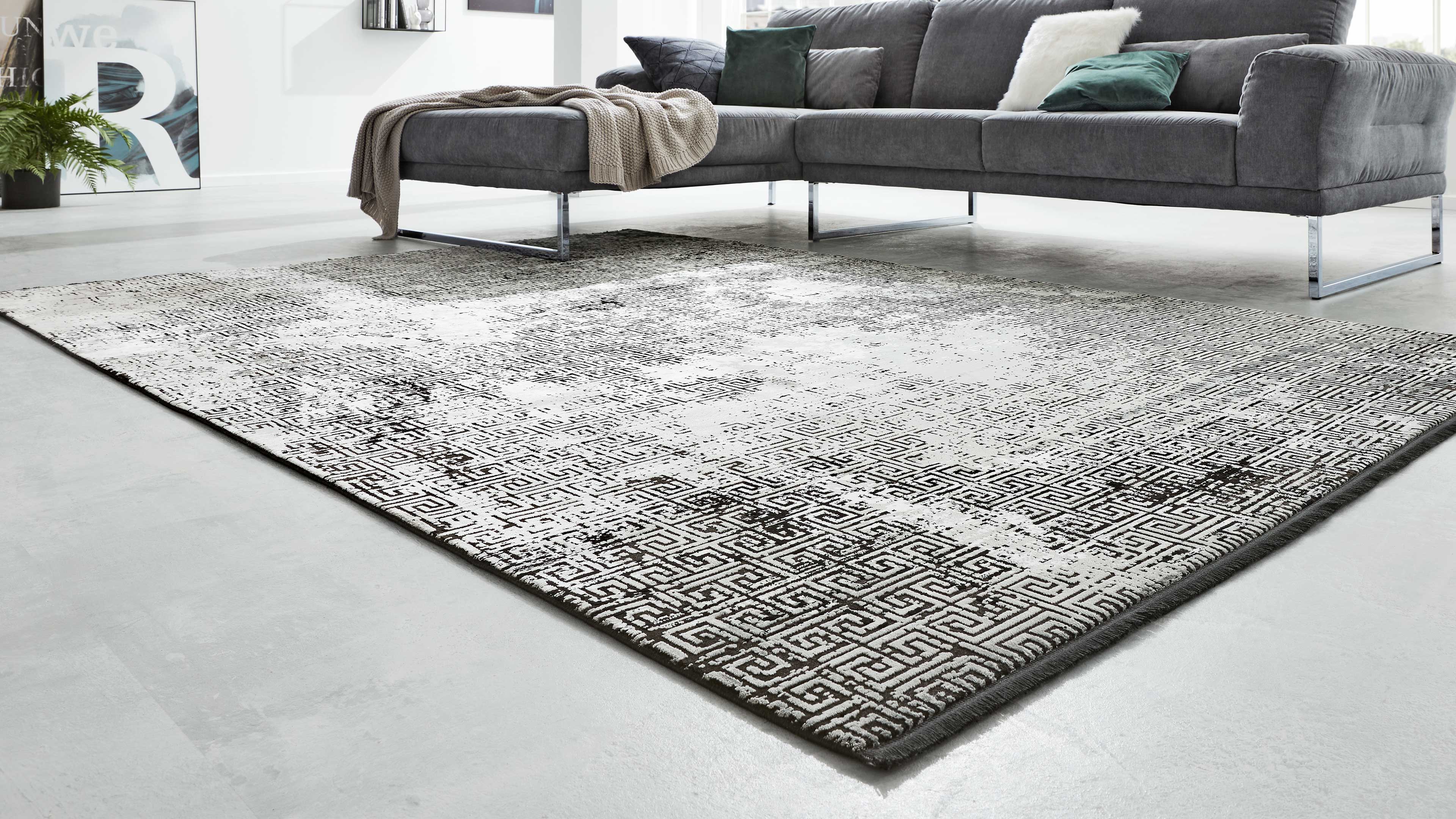 Großer rechteckiger Teppich mit weiß-dunkelgrauem geometrischem Muster auf hellem Boden vor einer dunklen Eckcouch mit Metallbeinen.