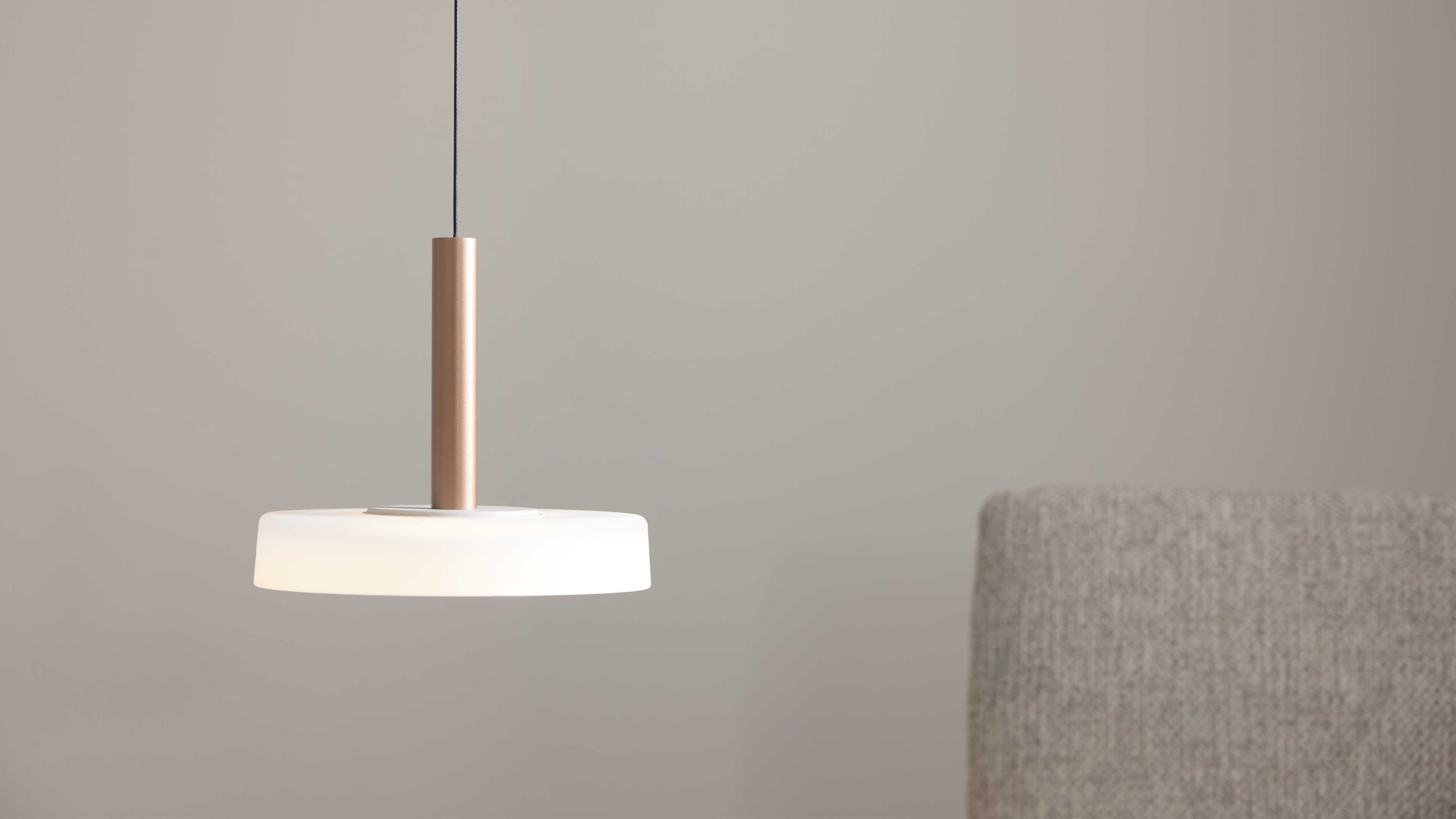 Hängende Pendelleuchte mit weißem Rundschirm und bronzefarbenem Zylinder am schwarzen Kabel, rechts ein grauer Stoffrücken.