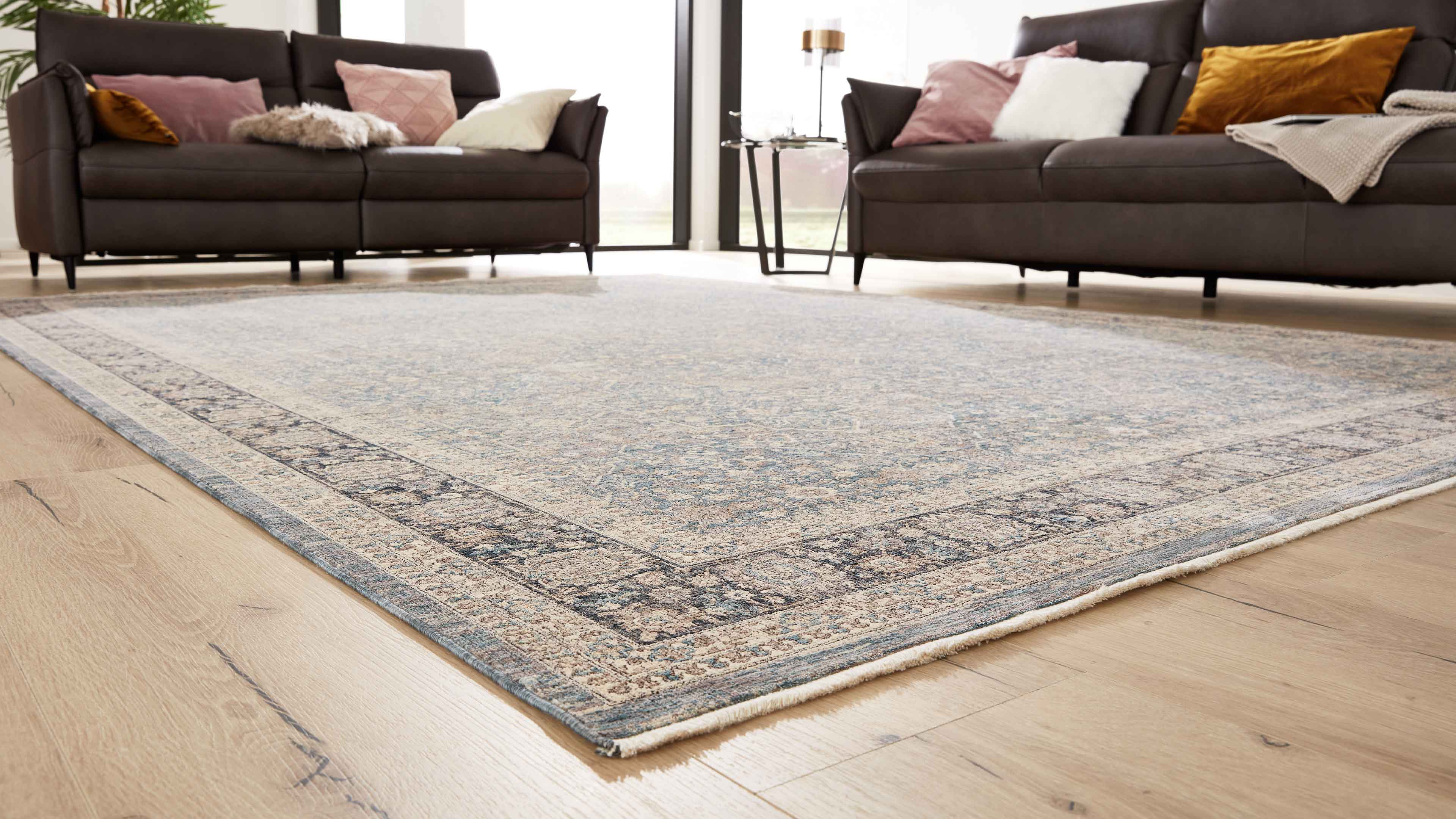 Großer hellblau-beiger Teppich mit Muster auf Holzboden, dahinter zwei dunkelbraune Sofas mit Kissen und Beistelltisch.