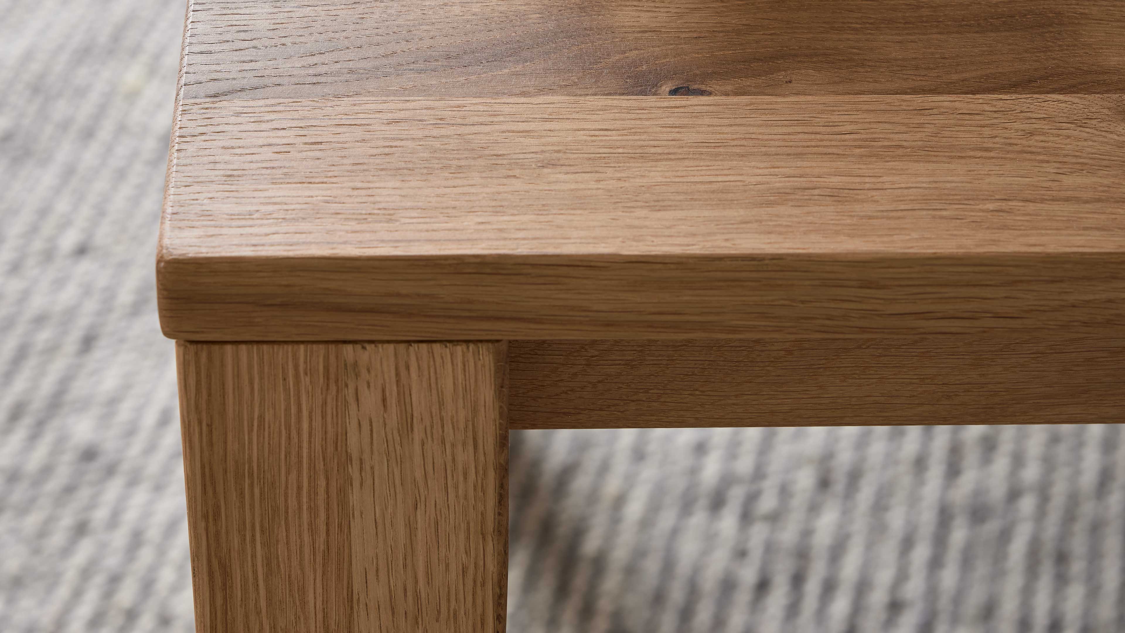 Eckansicht einer hellbraunen Holz-Tischplatte mit sichtbarer Maserung und eckigem Tischbein, im Hintergrund ein grauer Teppich.