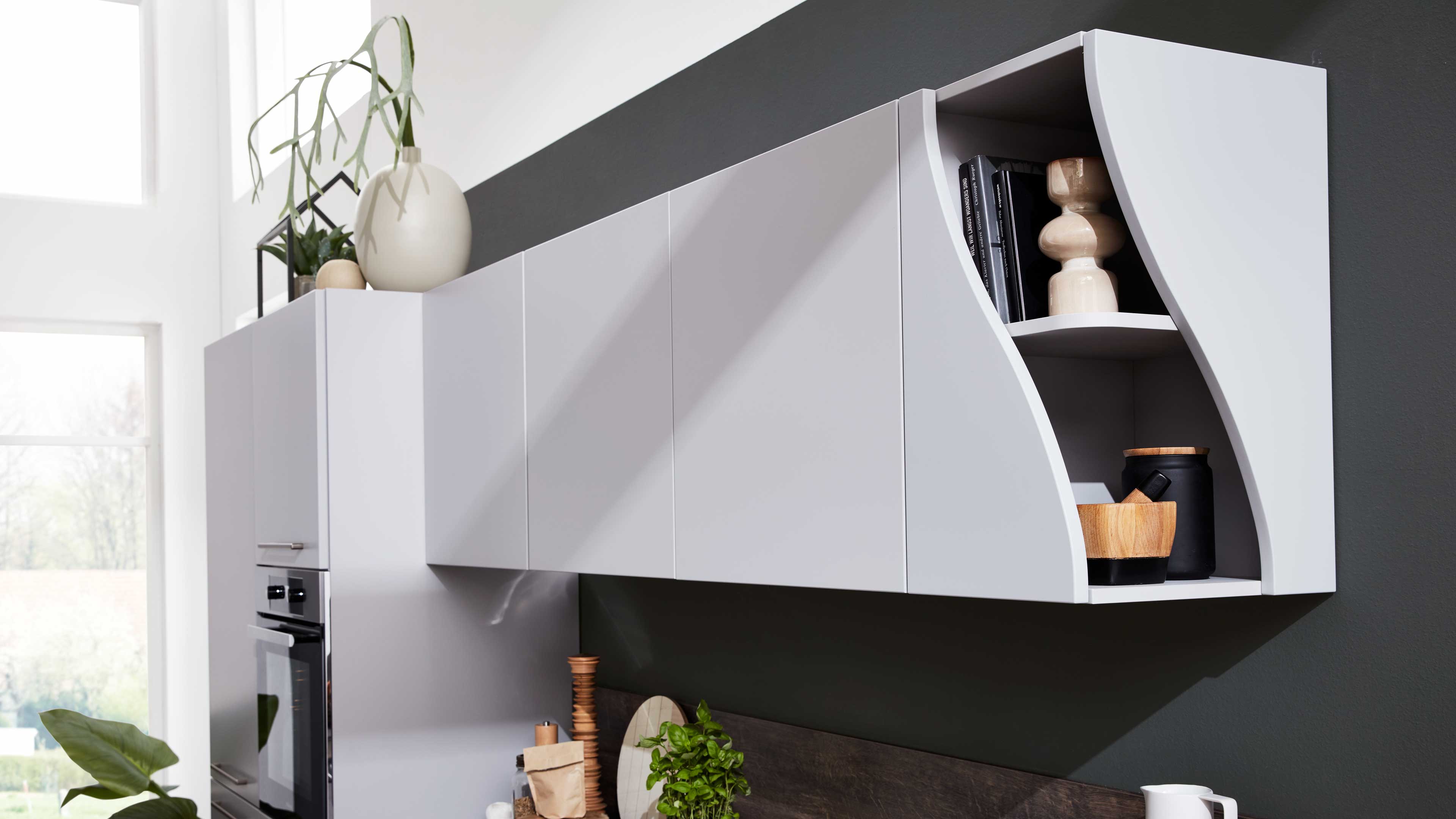 Hängender weißer Küchenoberschrank mit geschlossenen Fronten und rechts offener, geschwungener Regalnische mit Büchern und Deko.