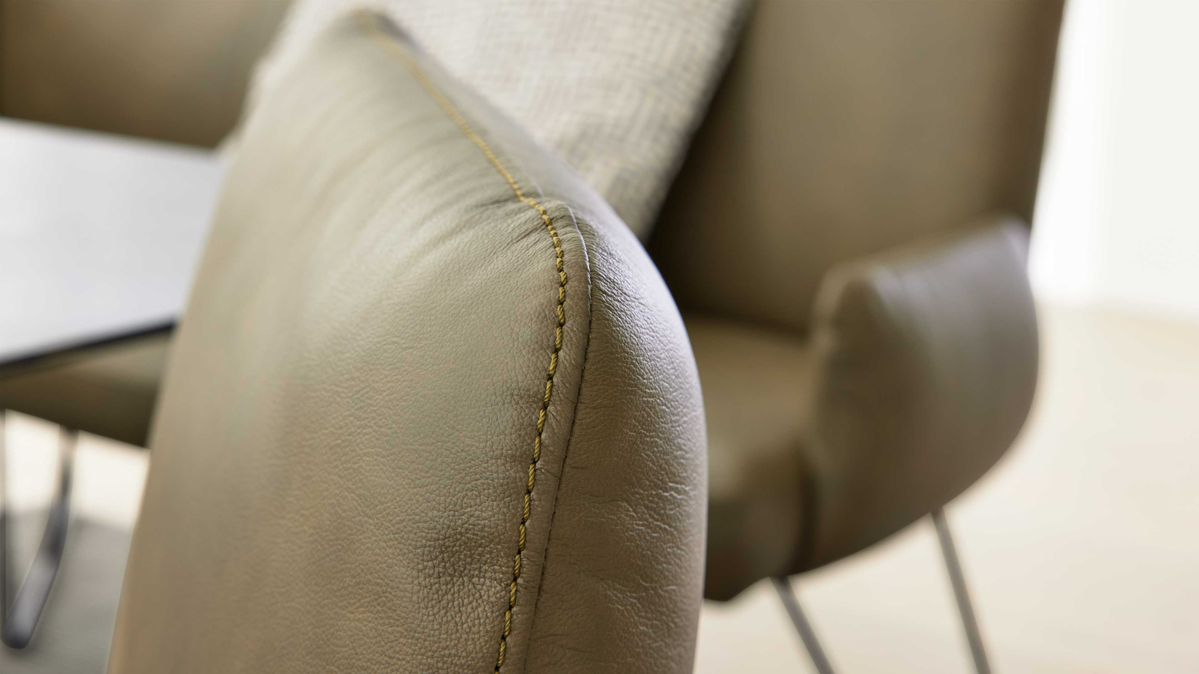 Nahaufnahme der Rückenlehne eines beigen Lederstuhls mit sichtbarer gelber Naht, unscharfer zweiter Stuhl und Metallbeine im Hintergrund.