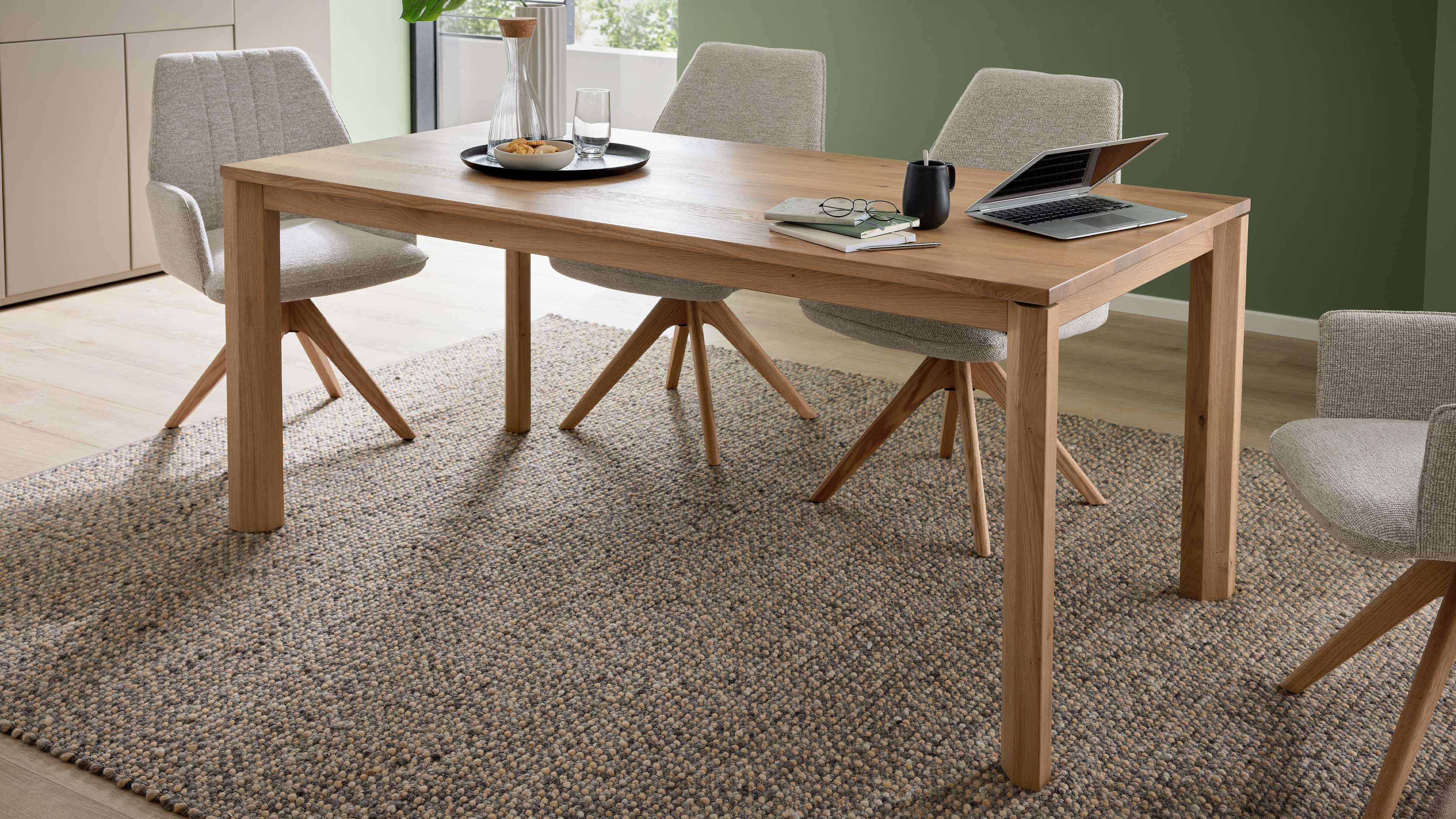 Rechteckiger Esstisch aus hellem Holz mit vier grauen, gepolsterten Stühlen auf Holzsternen, Laptop und Geschirr.