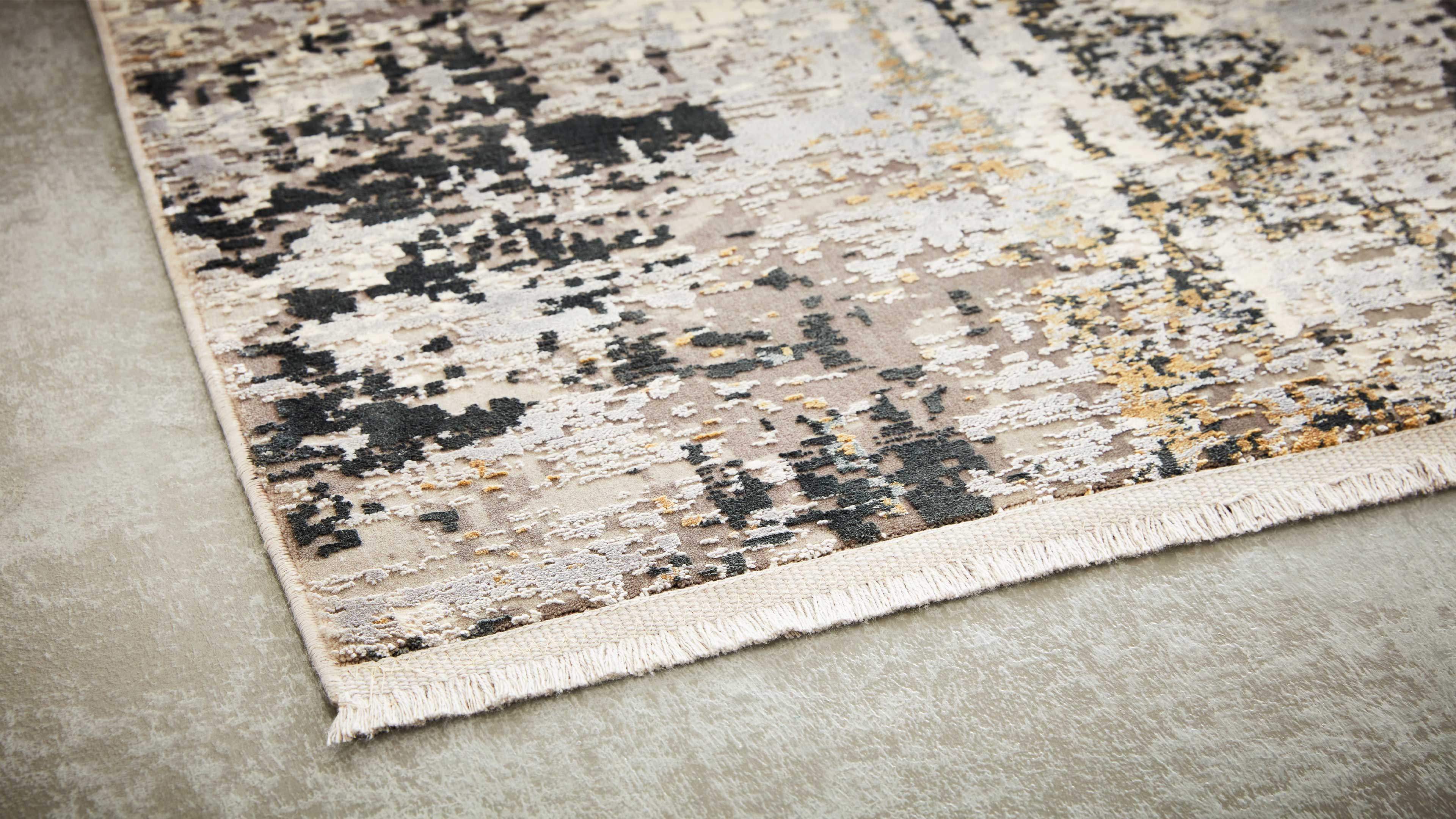 Eckansicht eines Teppichs mit beige-grauem, abstrakt gesprenkeltem Muster, schwarzen und goldenen Akzenten und Fransen auf hellem Boden.