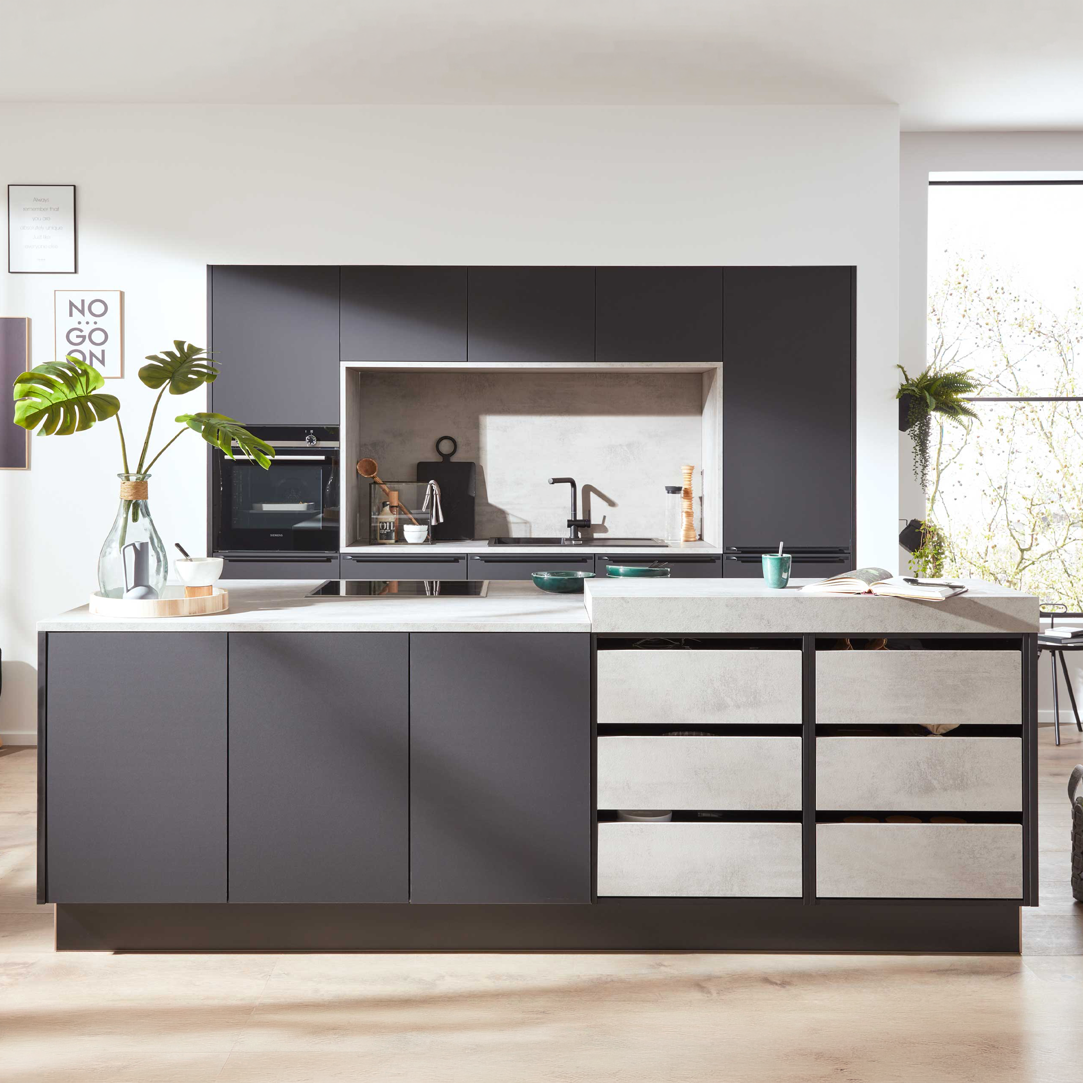 Moderne Küche mit großer dunkelgrauer Insel und Beton-Optik-Arbeitsplatte, Monstera-Vase, eingelassenem Spülbereich und großen Fenstern.