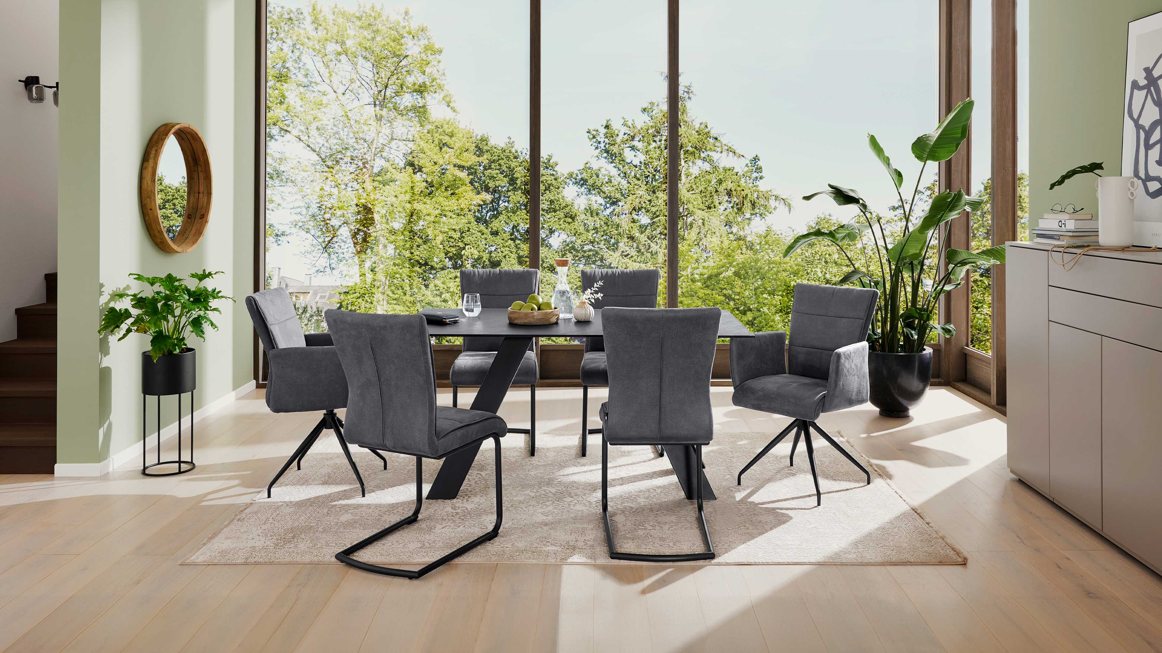 Esstisch mit rechteckiger dunkler Tischplatte und sechs grauen Polsterstühlen auf hellem Teppich vor großen Fensterfronten.