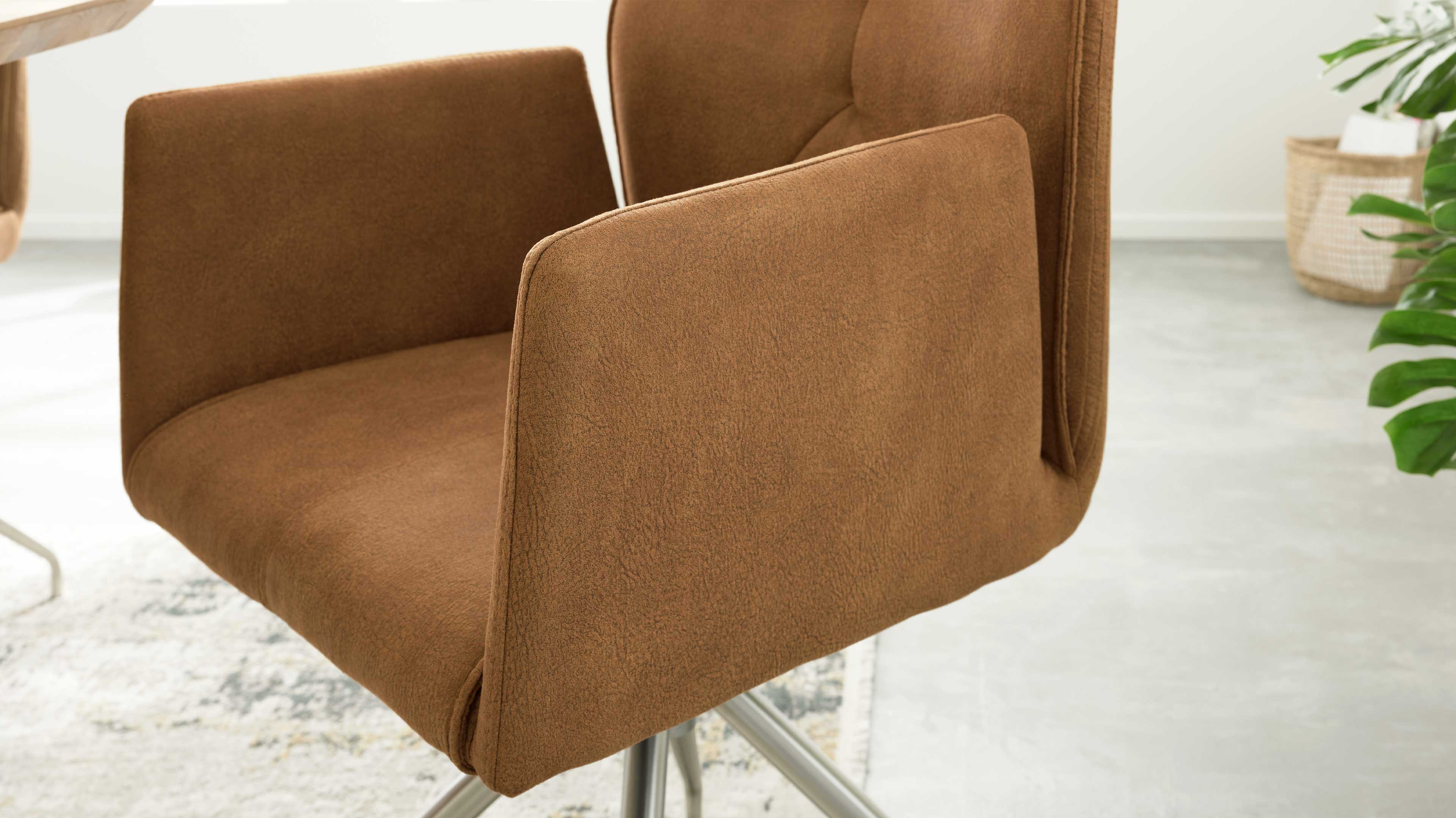 Braun bezogener Drehstuhl mit breiten Armlehnen und sichtbarem silbernem Metallkreuzfuß, Teppichkante, Pflanze im Hintergrund.