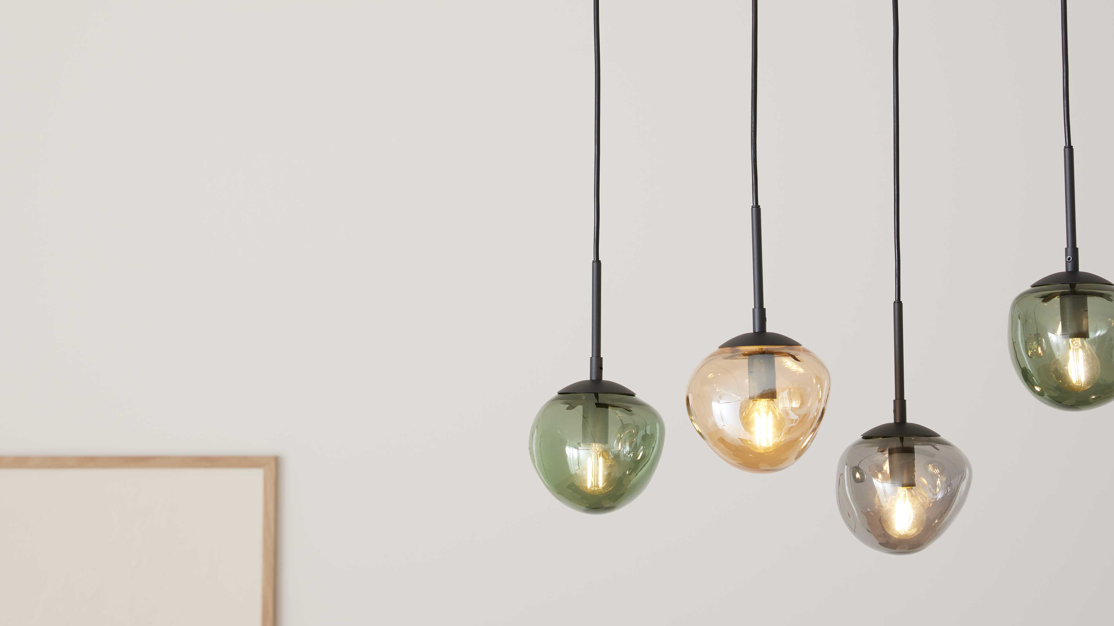 Vier hängende Glaspendelleuchten in Grün, Bernstein und Rauchglas mit schwarzen Kabeln und sichtbaren Leuchtmitteln vor heller Wand.