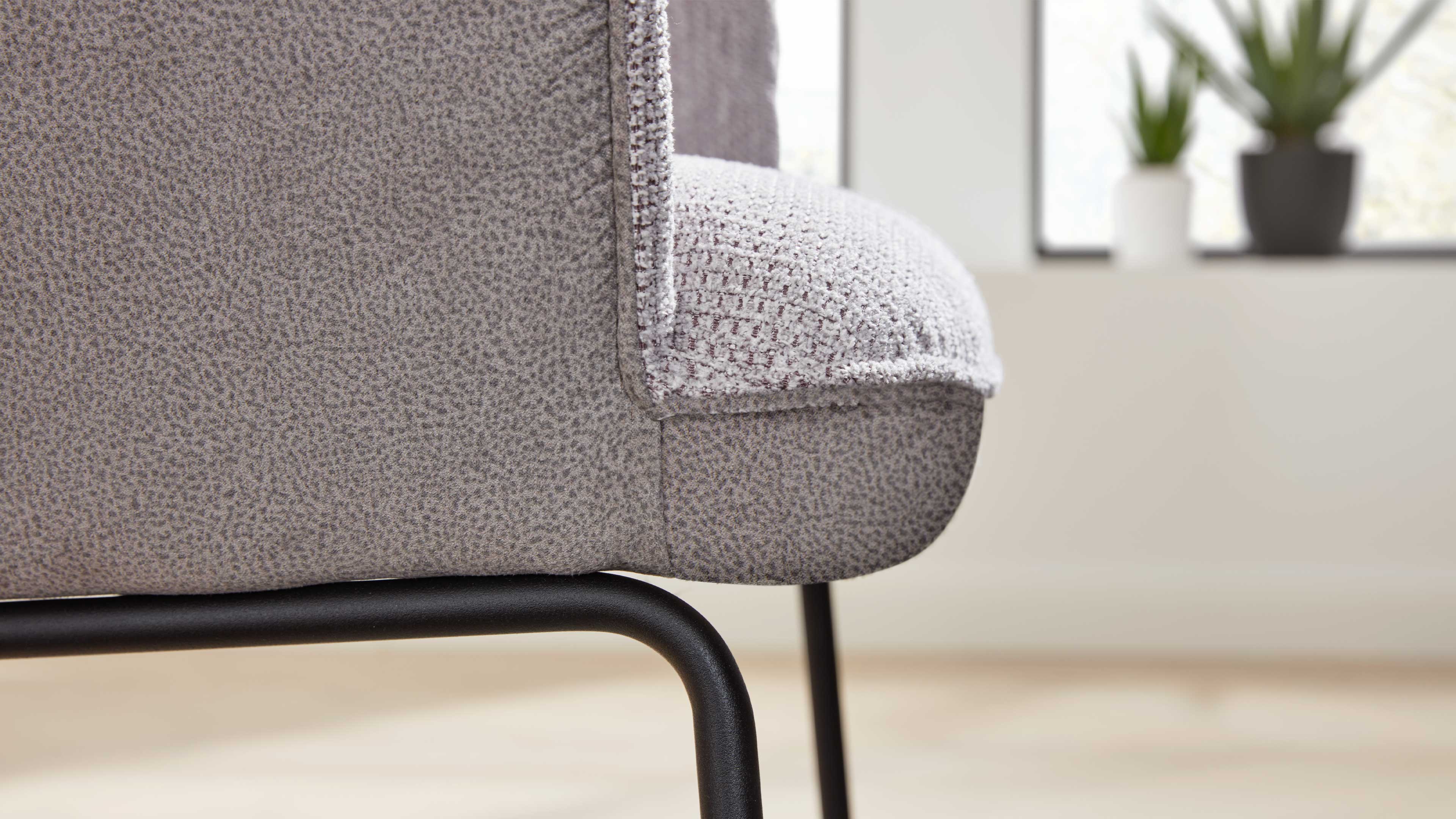 Nahaufnahme einer grauen, strukturierten Sofakante mit Polsterauflage und schwarzem Rundrohrgestell, im Hintergrund Pflanzen unscharf.