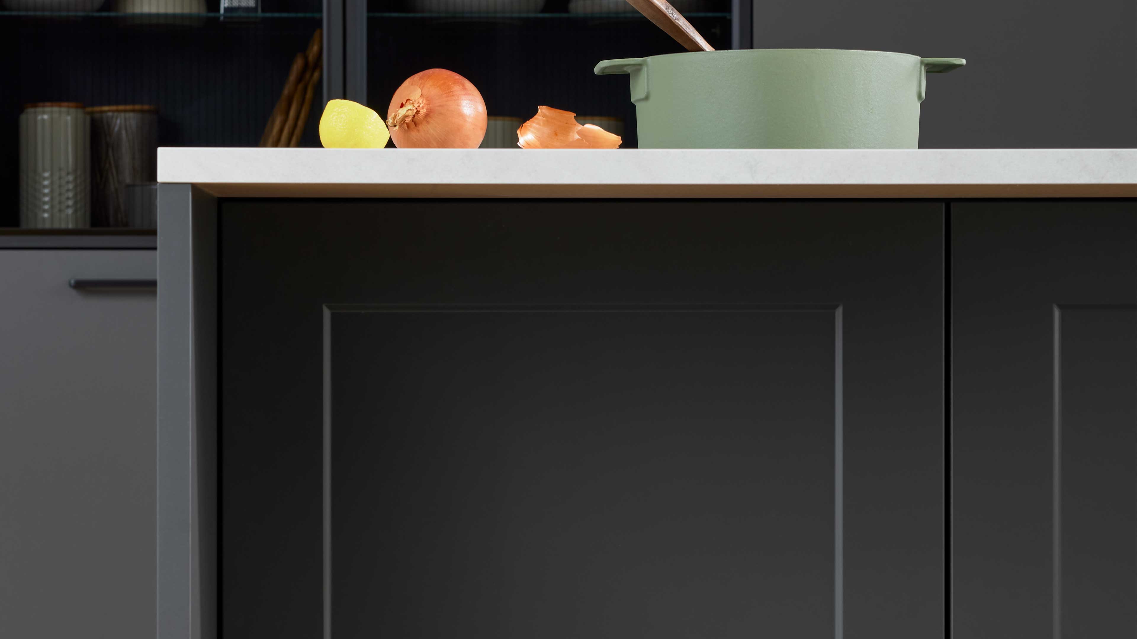 Dunkle Küchenunterschränke mit weißer Arbeitsplatte, darauf Zwiebel, Zitronenhälfte, Zwiebelschale und grüner Topf mit Holzlöffel.