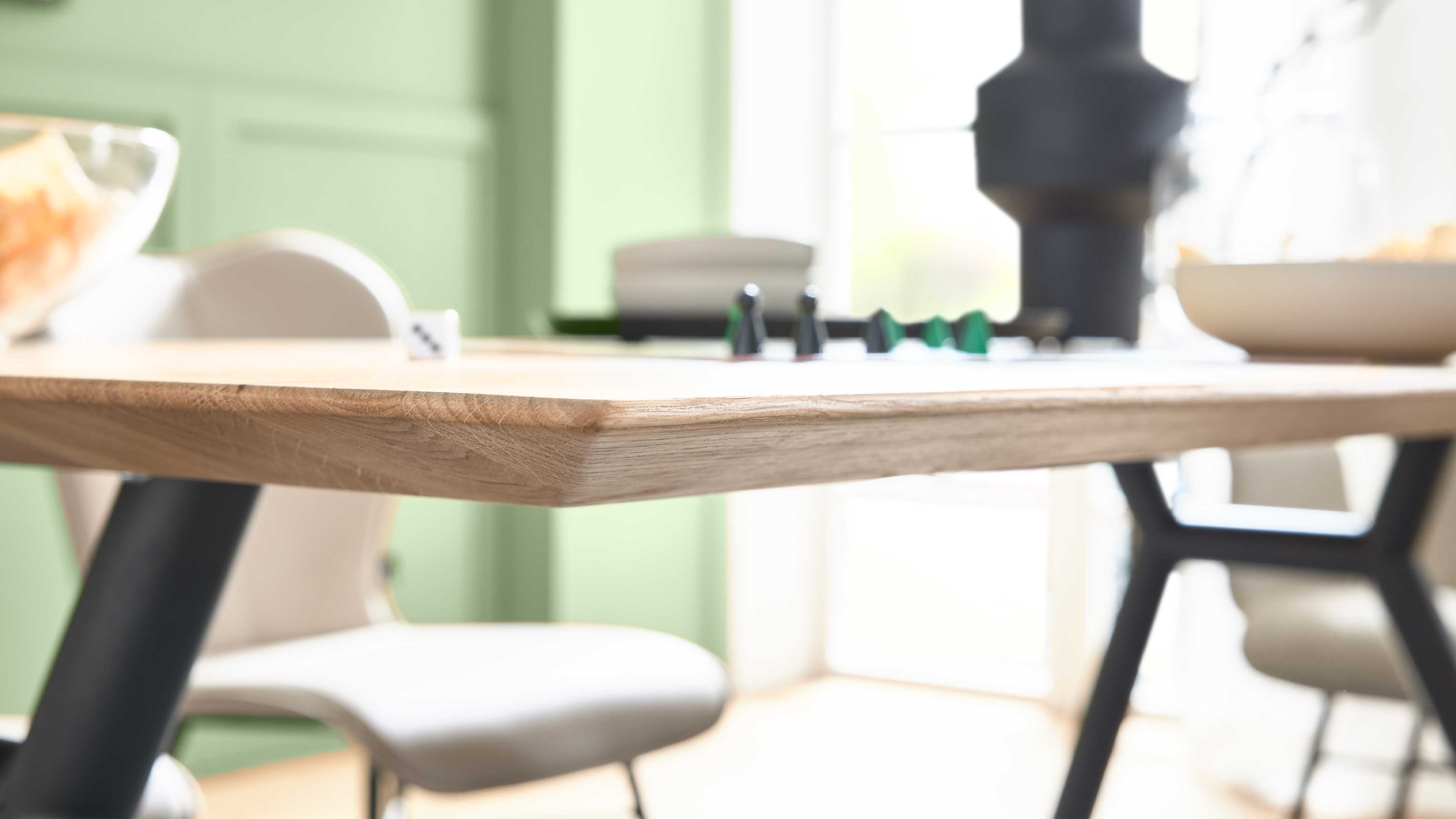 Eckansicht einer Holztischplatte mit abgerundeter Kante und schwarzen schrägen Metallbeinen, im Hintergrund Stuhl und Spielsteine.