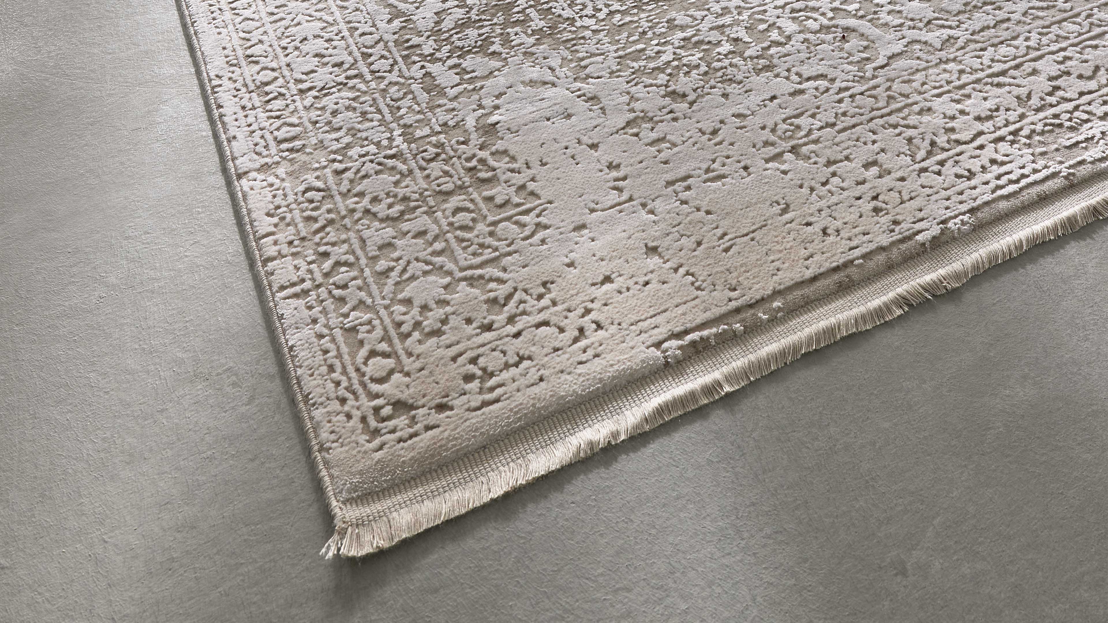 Heller beiger Teppich mit floralen und geometrischen Reliefmustern, gesäumte Fransen an der Ecke, liegt auf grauem Boden.