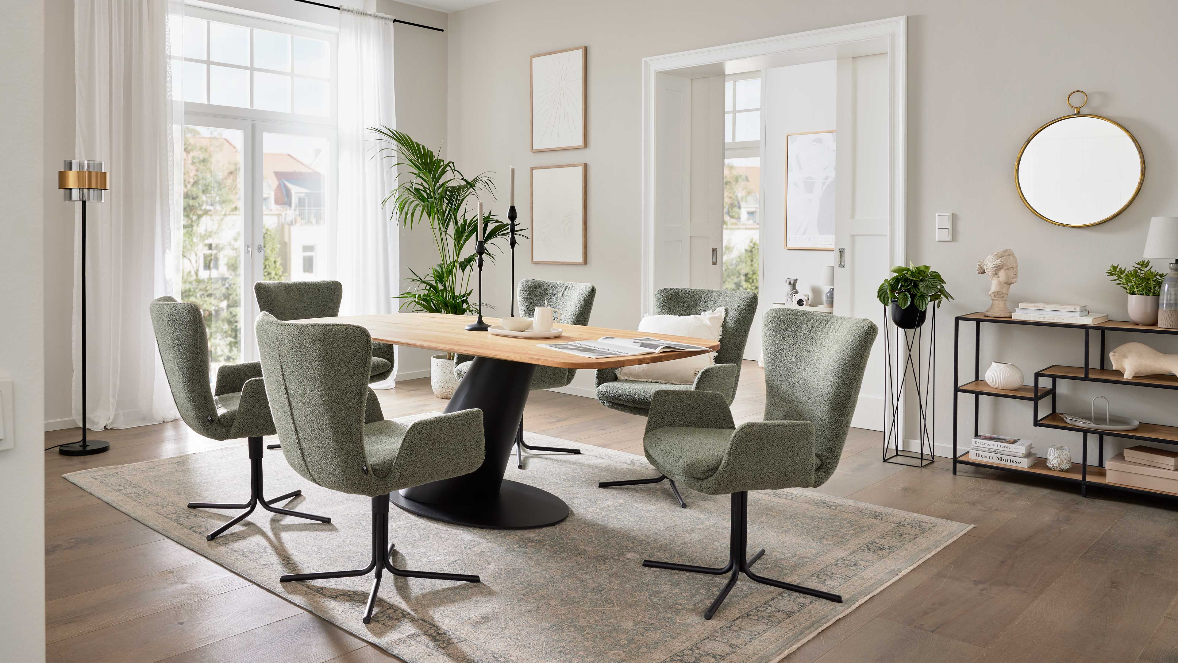 Ovaler Holztisch mit schwarzem Metallfuß und sechs grünen, gepolsterten Drehstühlen auf Teppich, daneben Regal, Spiegel und Pflanzen.