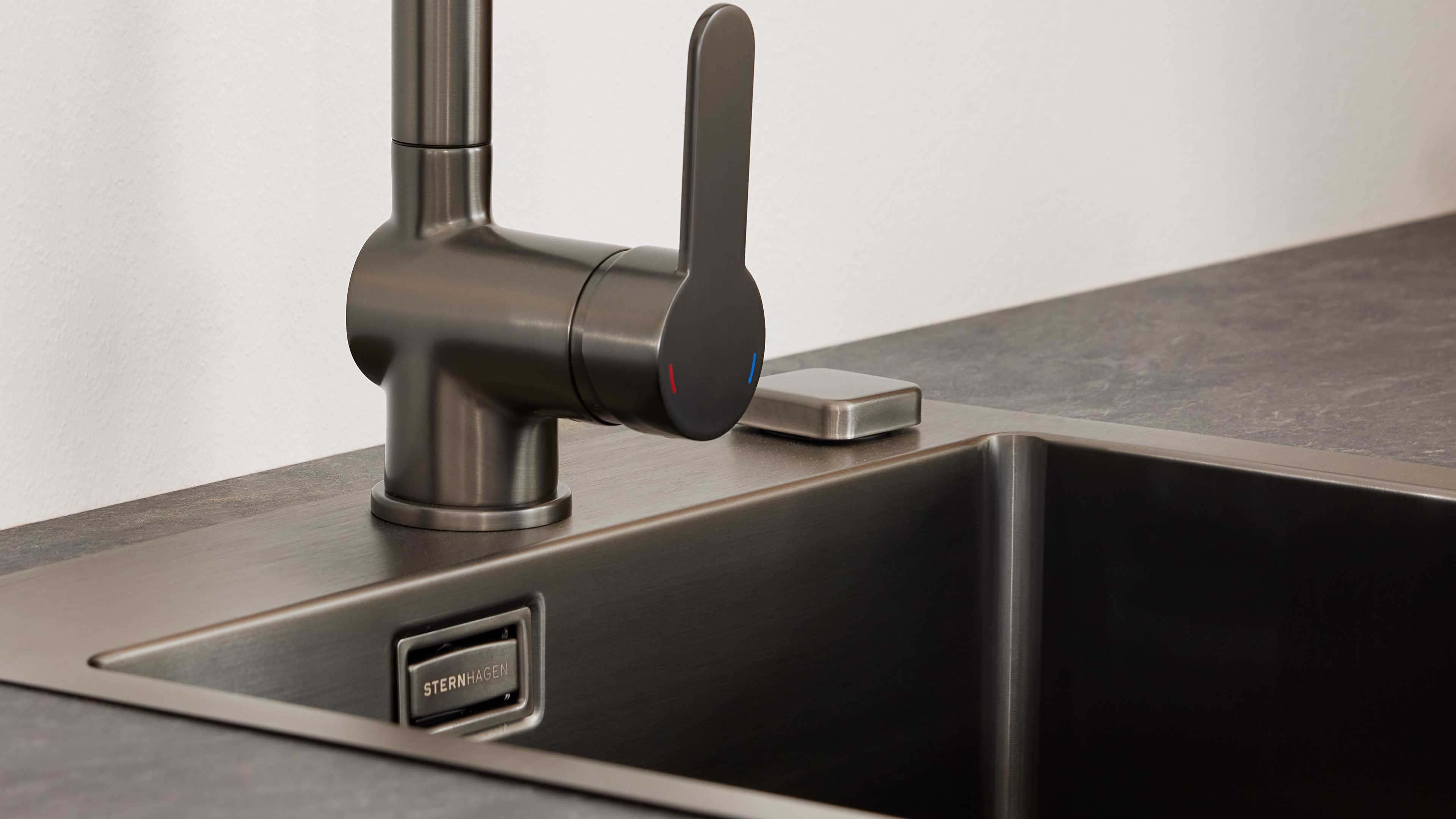 Dunkelgrauer gebürsteter Einhebelmischer auf Arbeitsplatte, daneben eine rechteckige Bedienplatte und schwarze Spüle mit Markenplakette.