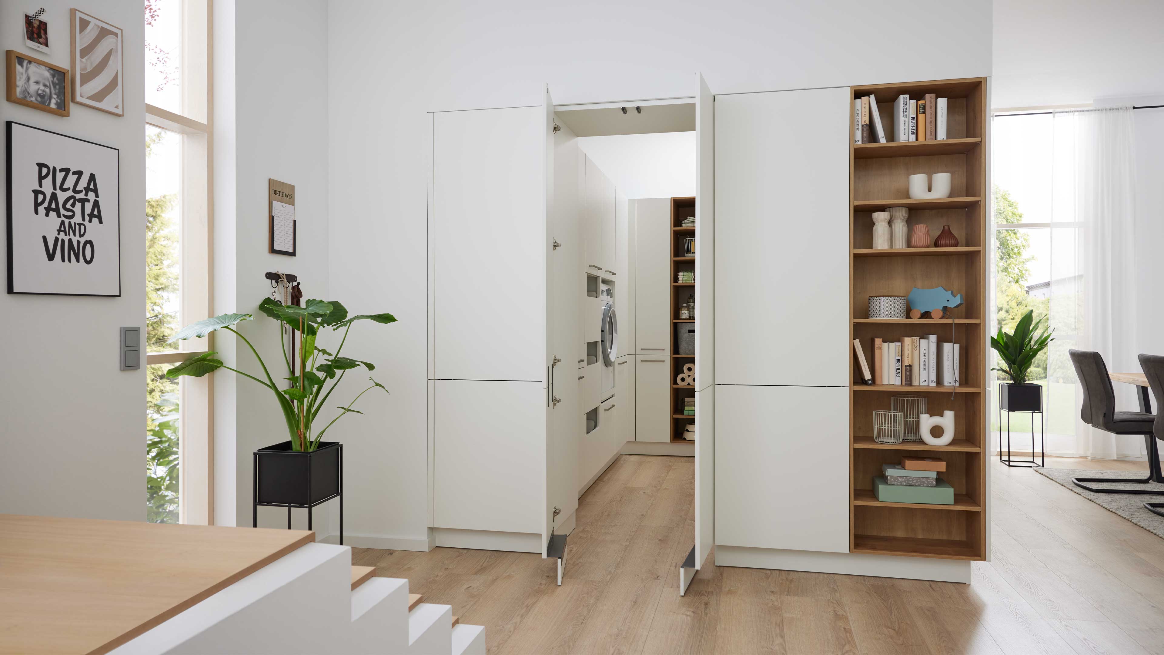 Weiße Einbau-Schrankwand mit geöffneten Mitteltüren, dahinter Hauswirtschaftsraum mit Waschmaschine, rechts offene Holzregale mit Büchern.