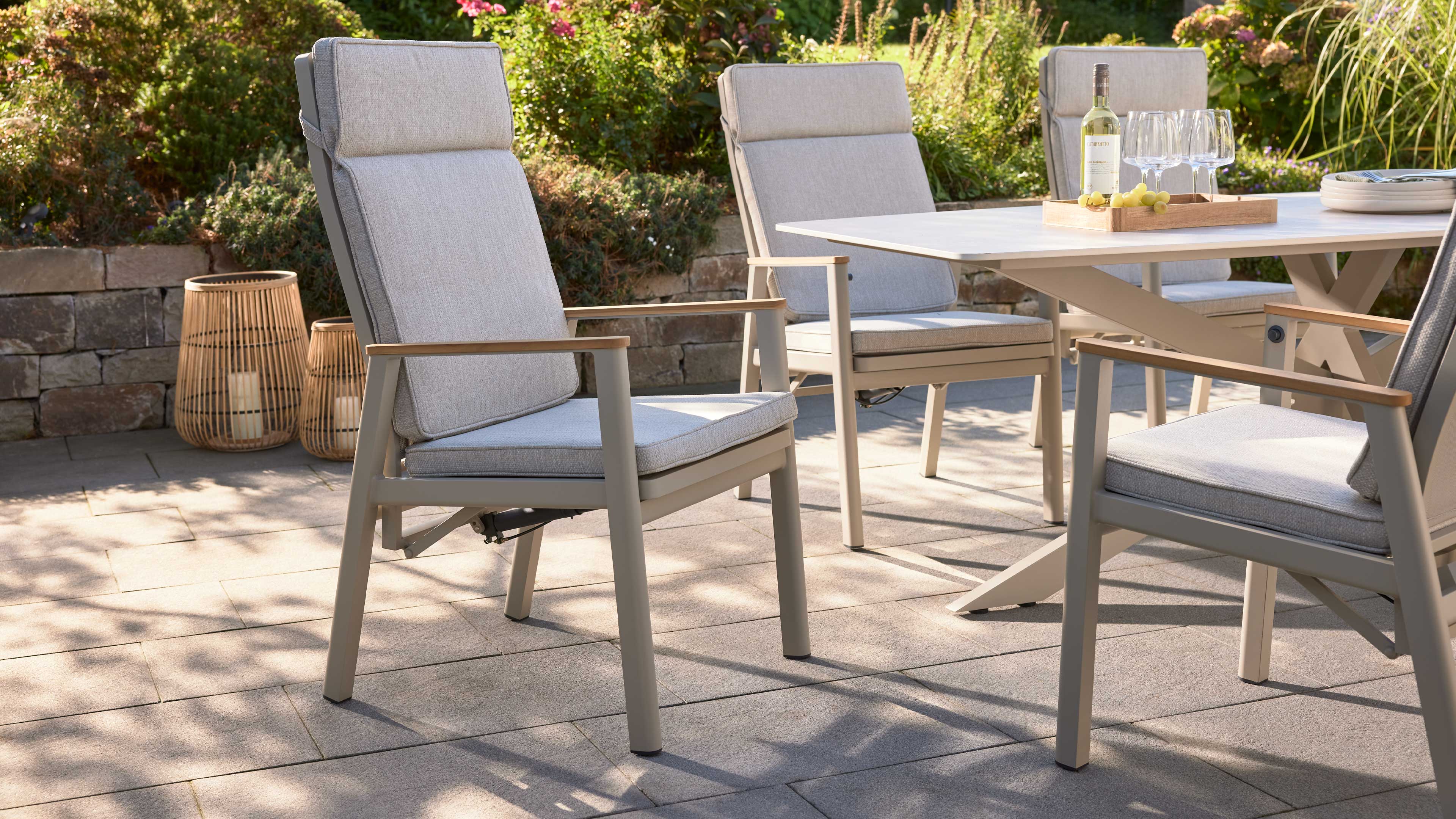 Vier gepolsterte Gartenstühle mit hellgrauen Kissen und beige-grauen Rahmen um einen hellen rechteckigen Tisch, auf Steinplatten.