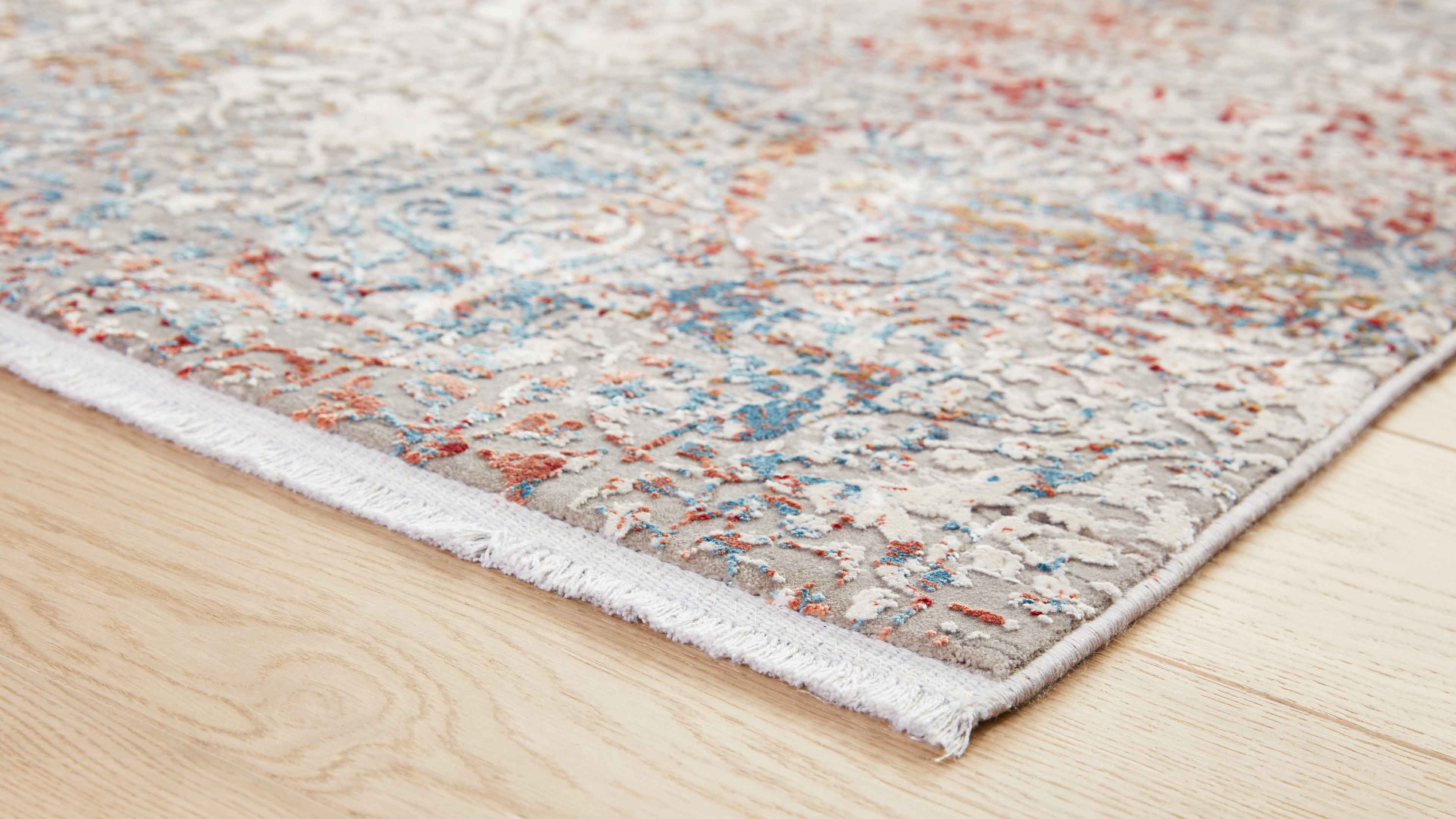Schräg fotografierte Teppichkante mit kurzem weißem Fransenbund, graubasigem Flor und blau-orangen Mustern auf hellem Holzboden.