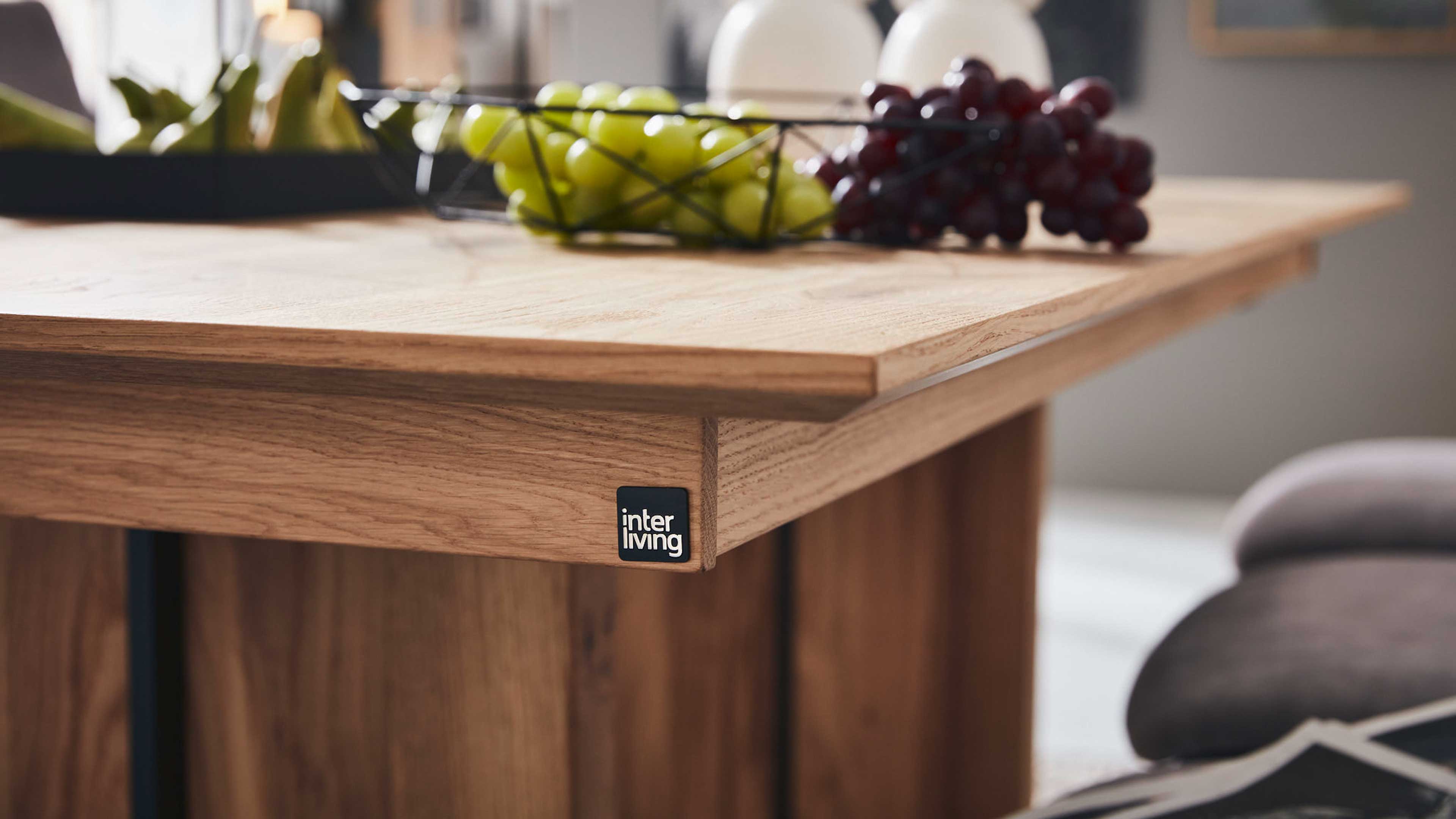 Eckansicht einer Holz-Tischplatte mit schwarzem inter living‑Label, darauf ein Drahtkorb mit grünen und roten Trauben.